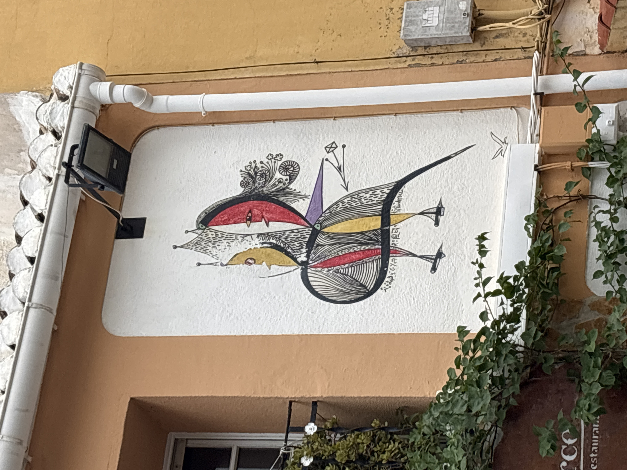 The image shows a mural on the exterior wall of a building. The mural is a stylized depiction of a bird-like creature with human-like facial features and ornate headwear. The creature is rendered in black ink with accents of yellow and red. The background of the mural is a textured white surface. To the left of the mural, there is a window with a green vine growing around it. To the right of the mural, a white downspout runs vertically along the wall. Above the mural, a white gutter and a black light fixture are visible. The building's walls are painted in shades of orange and yellow. A small electrical box is mounted on the yellow wall to the right of the downspout. In the lower left corner of the image, part of a red sign with white text is visible, which appears to say "erce restaurant". The overall setting suggests an outdoor urban environment.
