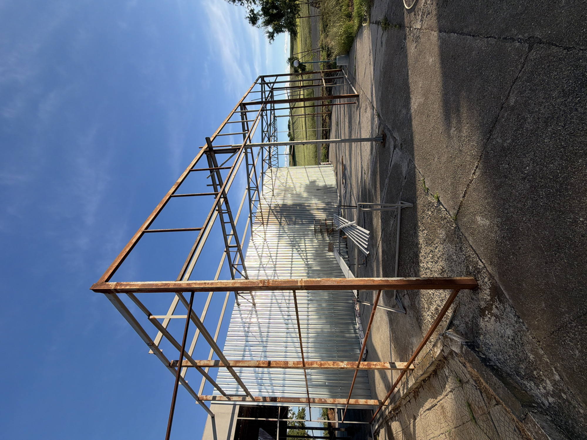 The image shows a metal framework structure, likely a greenhouse or similar agricultural building, standing outdoors. The metal is rusty in places. Behind the structure is a corrugated metal sheet partially covering the back. The ground is concrete, cracked and weathered. In the background, there is a grassy field and some trees. The sky is blue with some clouds. The location is Dabnik, Bulgaria.