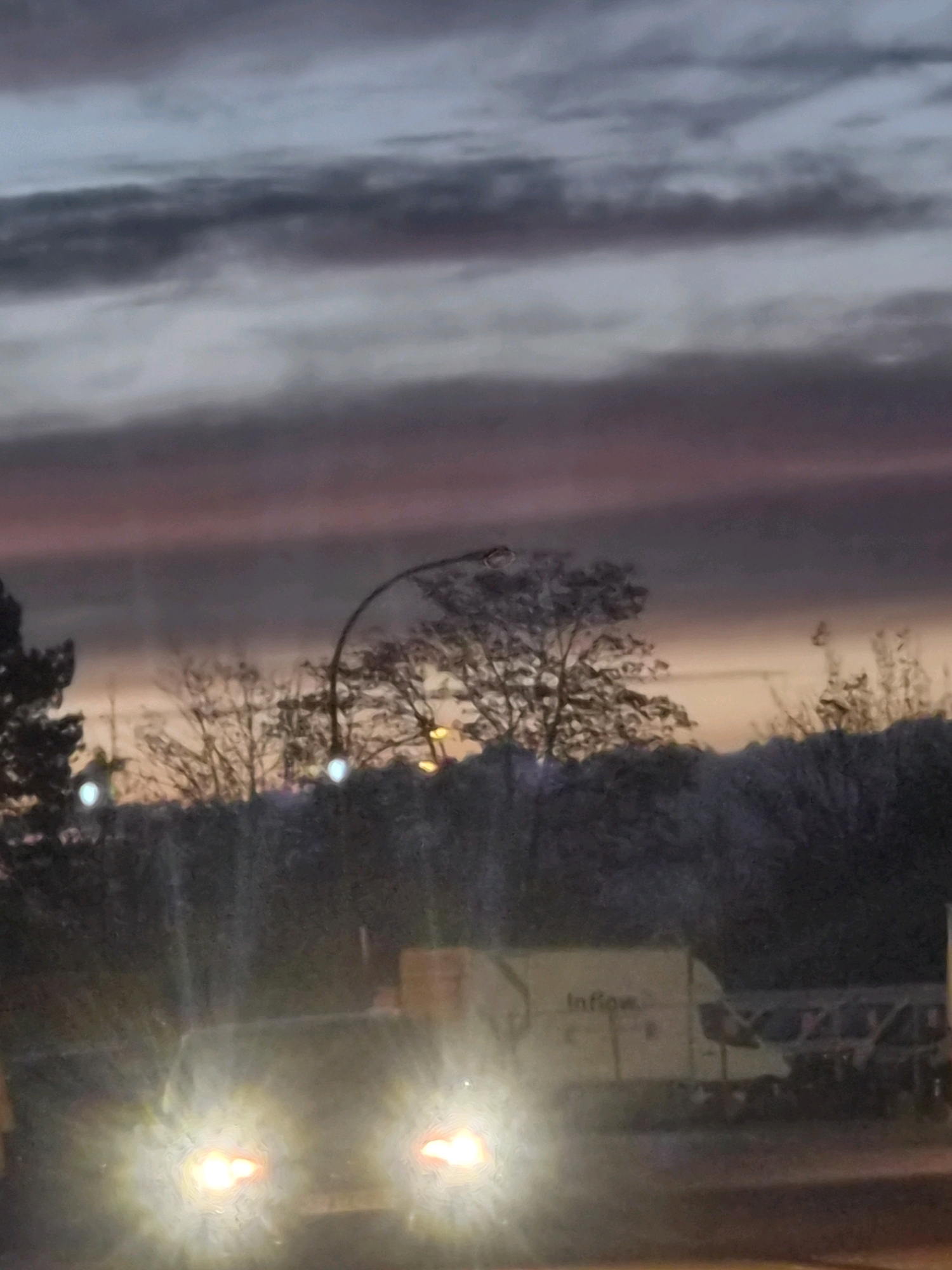 The image depicts a twilight scene possibly in Leudelange, Luxembourg, characterized by a soft gradient of colors in the sky, transitioning from dark grey clouds at the top to pink and orange hues near the horizon. Silhouetted trees and foliage form a dense line across the middle ground, with faint streetlights visible among them. In the foreground, the headlights of a vehicle are brightly illuminated, casting a glare on the image. Behind the headlights, a light-colored truck with the text "Inflow" visible on its side is parked. The overall impression is that of a dusk or early evening setting, with artificial light sources interacting with the natural dimness.