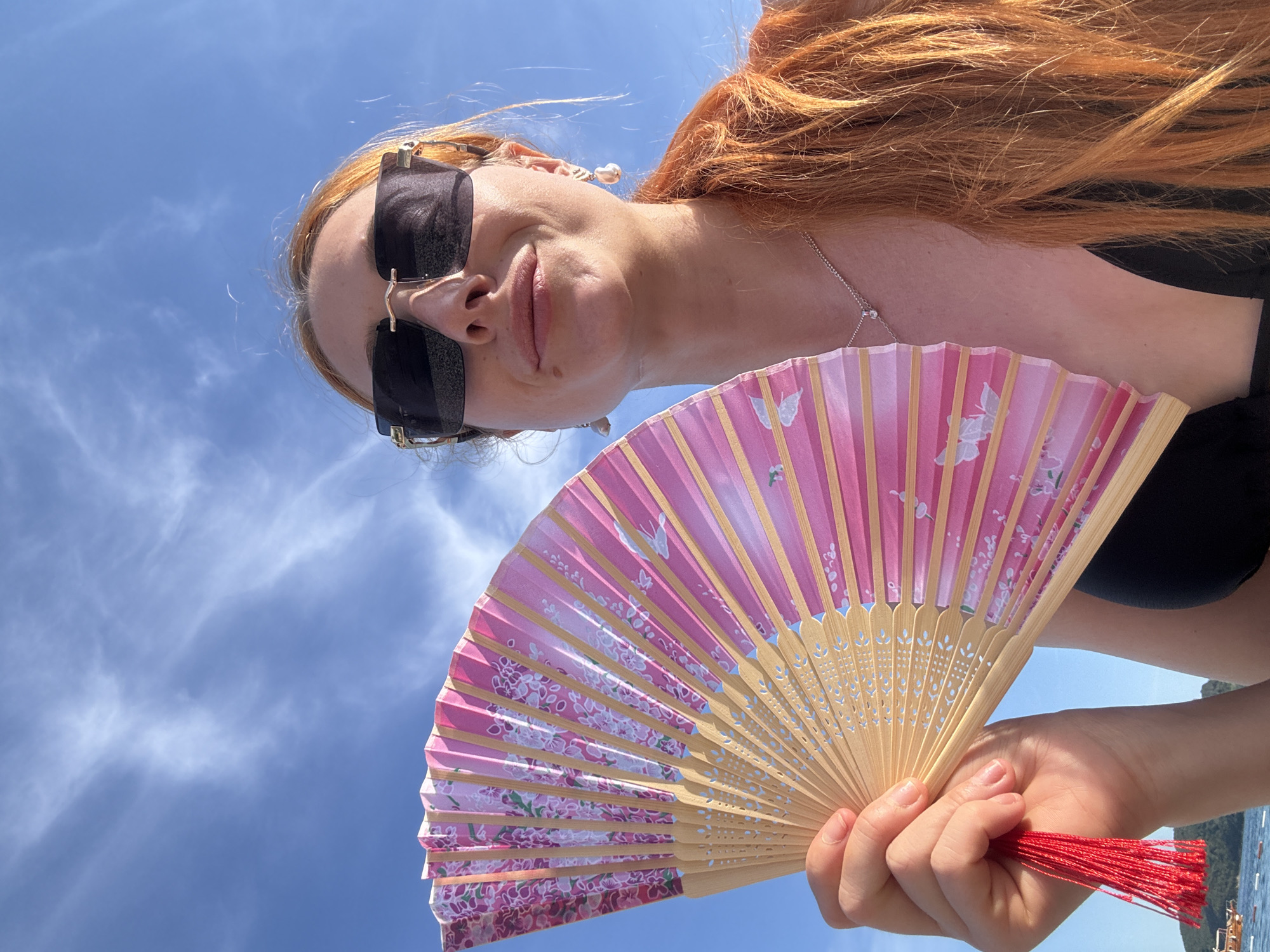A young woman with red hair is holding a pink, floral-patterned fan against a bright blue sky dotted with light clouds. She is wearing sunglasses and dangling earrings. The woman is wearing a black top, possibly a bikini top. A delicate silver necklace adorns her neck. In the background, there is a glimpse of a coastal area with trees and boats, suggesting a seaside location. The image appears to be taken from a low angle, looking up towards the sky and the woman. The setting appears to be in or near Budva, Montenegro, given the coastal scenery.