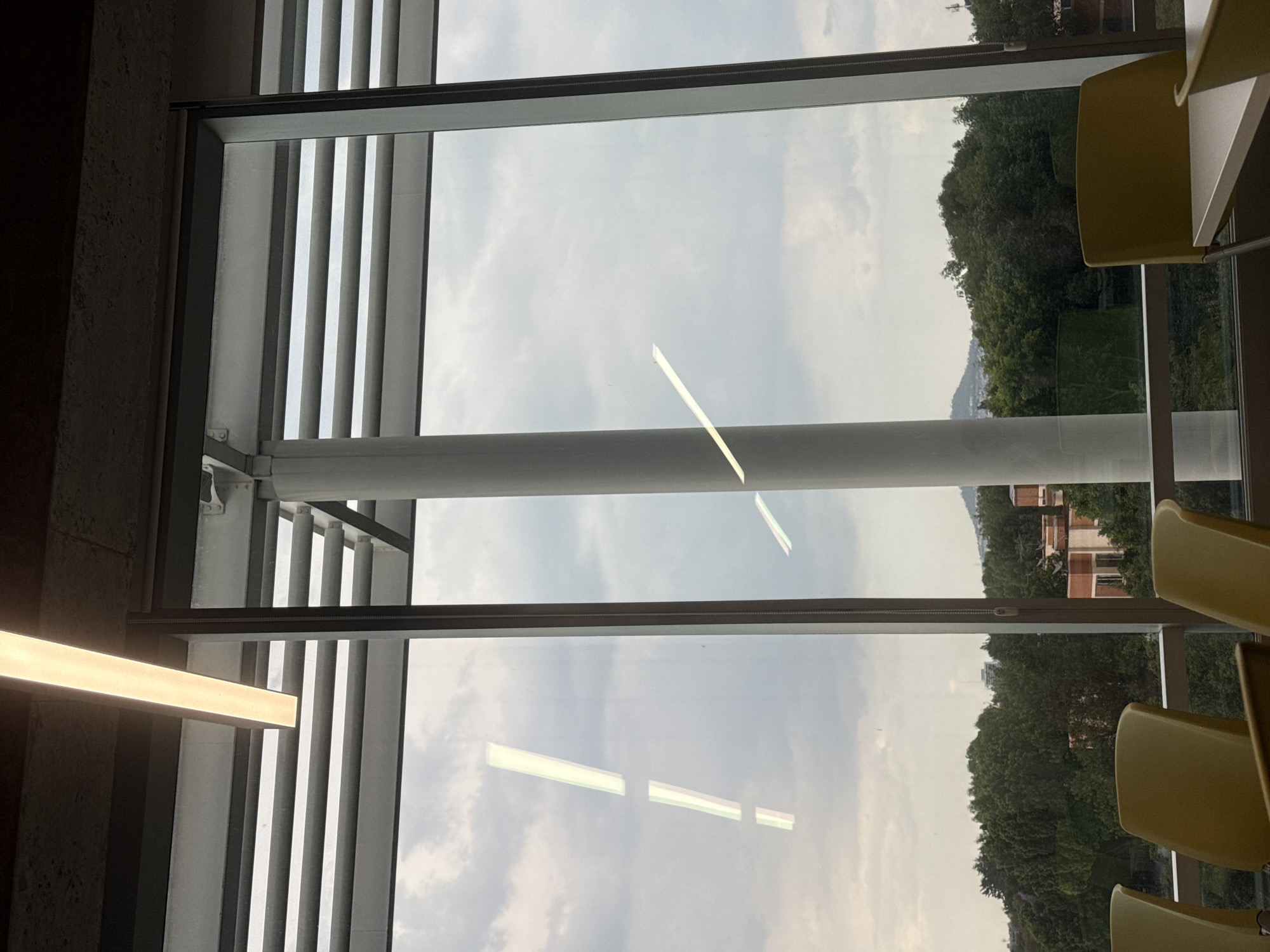 The image shows a view through a window with horizontal blinds, likely in a building, possibly a cafeteria or study area, as suggested by the presence of yellow chairs and a table. Outside, trees and a building can be seen, suggesting an urban or suburban location. The sky is cloudy, casting a slightly overcast light onto the scene. Reflections are visible on the glass of the window. There is no specific information in the image to indicate it is in Mexico City, Mexico.