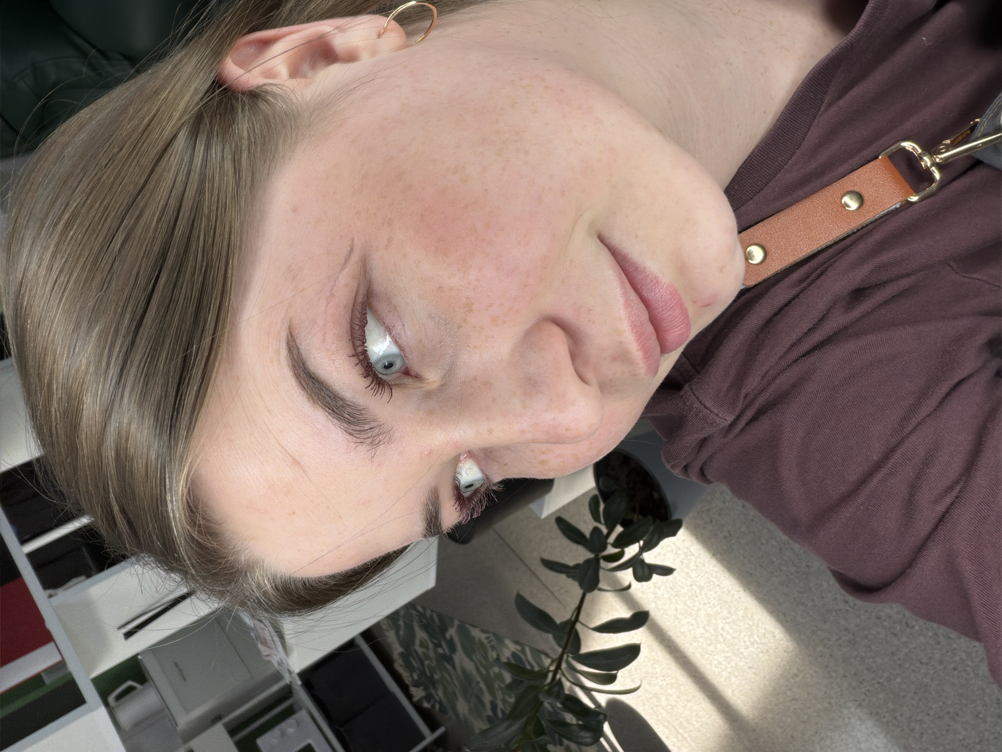 A close-up, angled portrait shows a woman with fair skin and light brown hair, noticeable freckles, and light blue eyes. She is wearing a gold hoop earring in her left ear and a brown shirt or top. A tan-colored strap with gold hardware is visible near her shoulder. The background includes part of a shelf unit with various storage boxes and items, a houseplant with green leaves, and a light-colored carpet with sunlight. The location in the city of Chaikovsky, Russia, is not definitively discernible from the visual information provided.