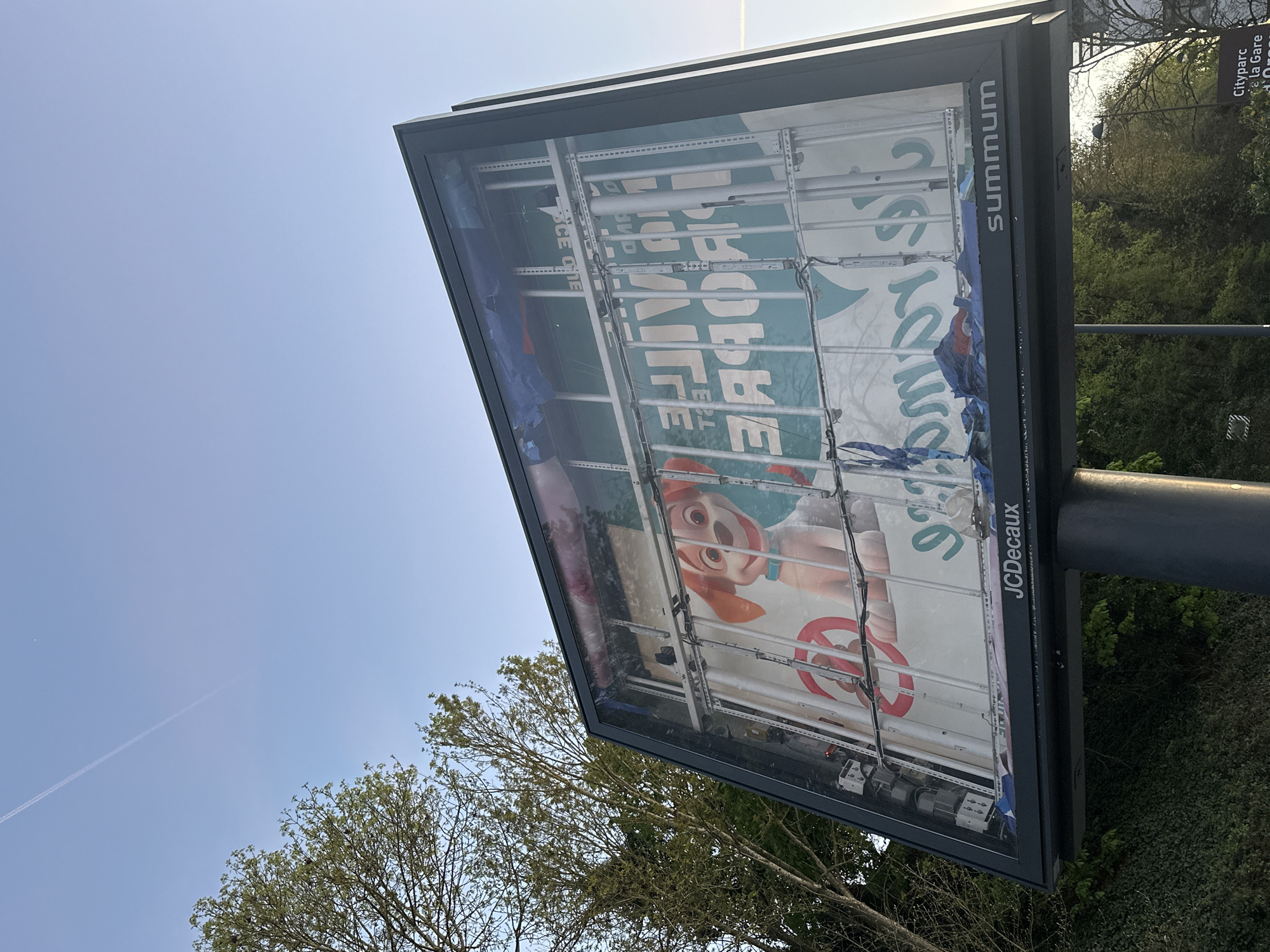 A low-angle shot reveals a billboard advertisement in Orsay, France, possibly located near the Cityparc and the Gare d'Orsay. The advertisement is partially torn and peeling, revealing glimpses of the underlying structure and previous posters. The remaining visible advertisement displays a cartoon dog character with an orange head and collar, holding what appears to be a red and white traffic sign. The word "LIFE" is prominently displayed in large, stylized letters, along with the phrase "HopE by." The billboard is branded with "Summum" on one side and "JCDecaux" on another. The background features a clear blue sky with a faint contrail, and foliage from nearby trees. The billboard is mounted on a dark metal pole.