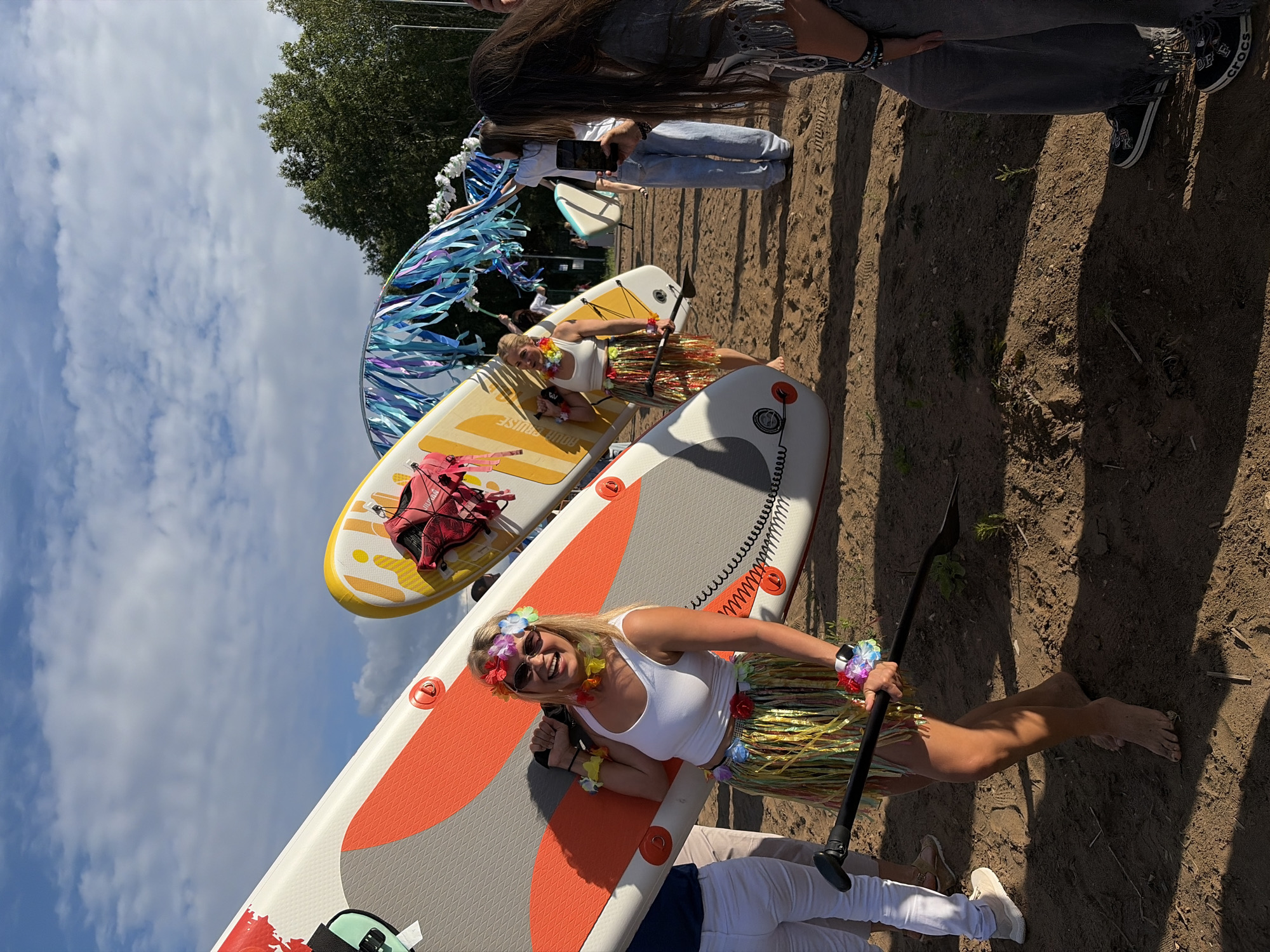 In Chaikovsky, Russia, a group of people are gathered on a sandy area, likely near a body of water, with several paddleboards present. The foreground features a woman wearing a white tank top, a colorful lei necklace, a flower crown, sunglasses, and a grass skirt. She is holding a paddleboard and a paddle. Behind her, another woman in a similar grass skirt and lei is standing near a yellow paddleboard. Further in the background, other individuals are visible, including someone holding a smartphone and capturing a photo. A decorative arch adorned with blue and purple streamers is also present in the scene. The sky is partly cloudy, and trees line the horizon. The overall atmosphere appears festive, possibly related to a water sports event or celebration.