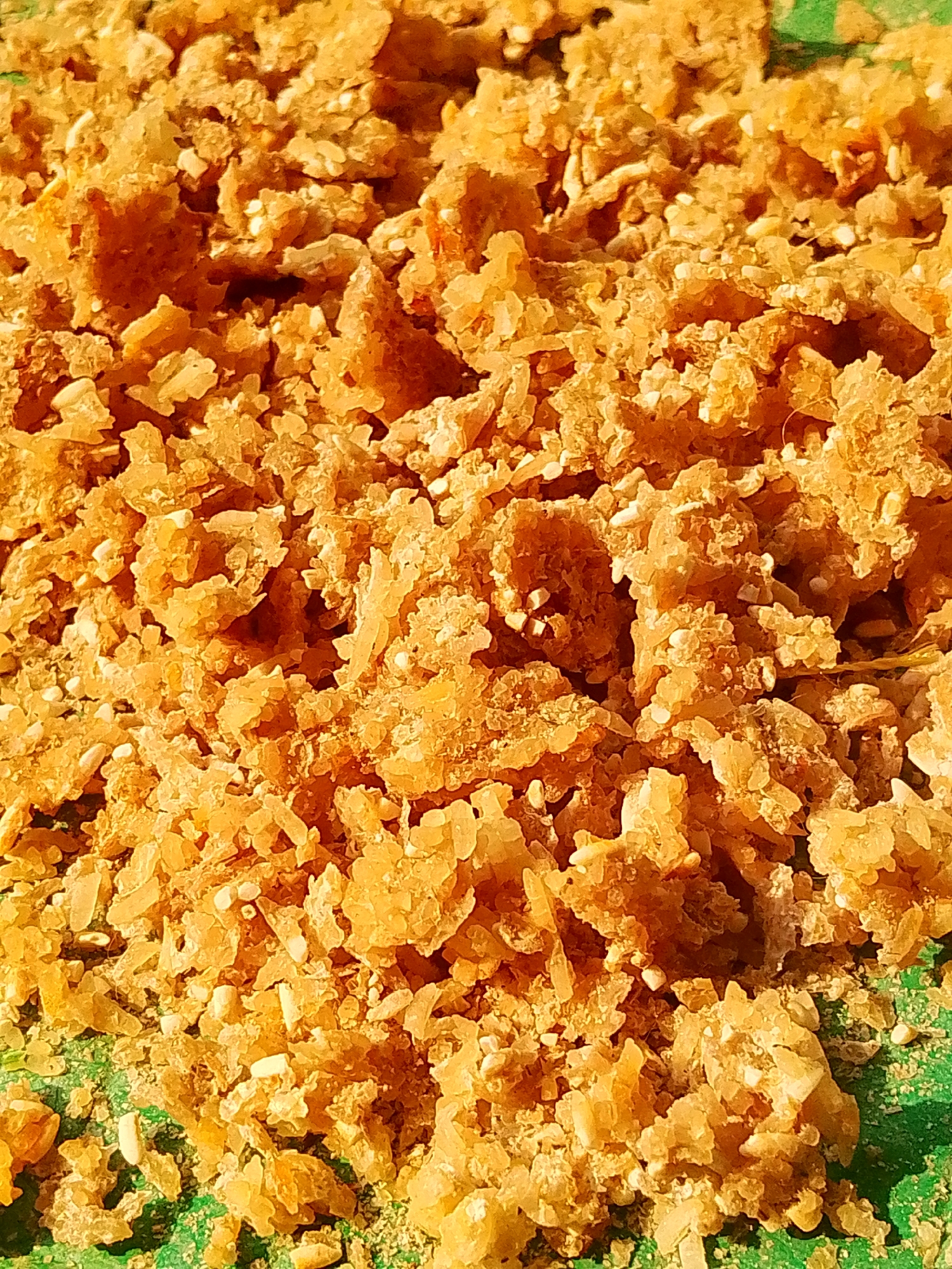 A close-up, overhead shot shows a pile of golden-brown, granular material, possibly crushed rice or puffed rice, mixed with small white seeds. The texture is rough and irregular, with some pieces appearing larger and more crystalline than others. The material is spread across a green surface, which appears to be a placemat or tray, partially visible at the bottom and left edges of the frame. The lighting suggests it is daytime, casting slight shadows and highlighting the texture of the food. This scene appears to be a food preparation area, potentially in a home or a small market stall in Barabai, Indonesia, given the context of the location. There are no people or discernible actions taking place, only the static arrangement of the food item.