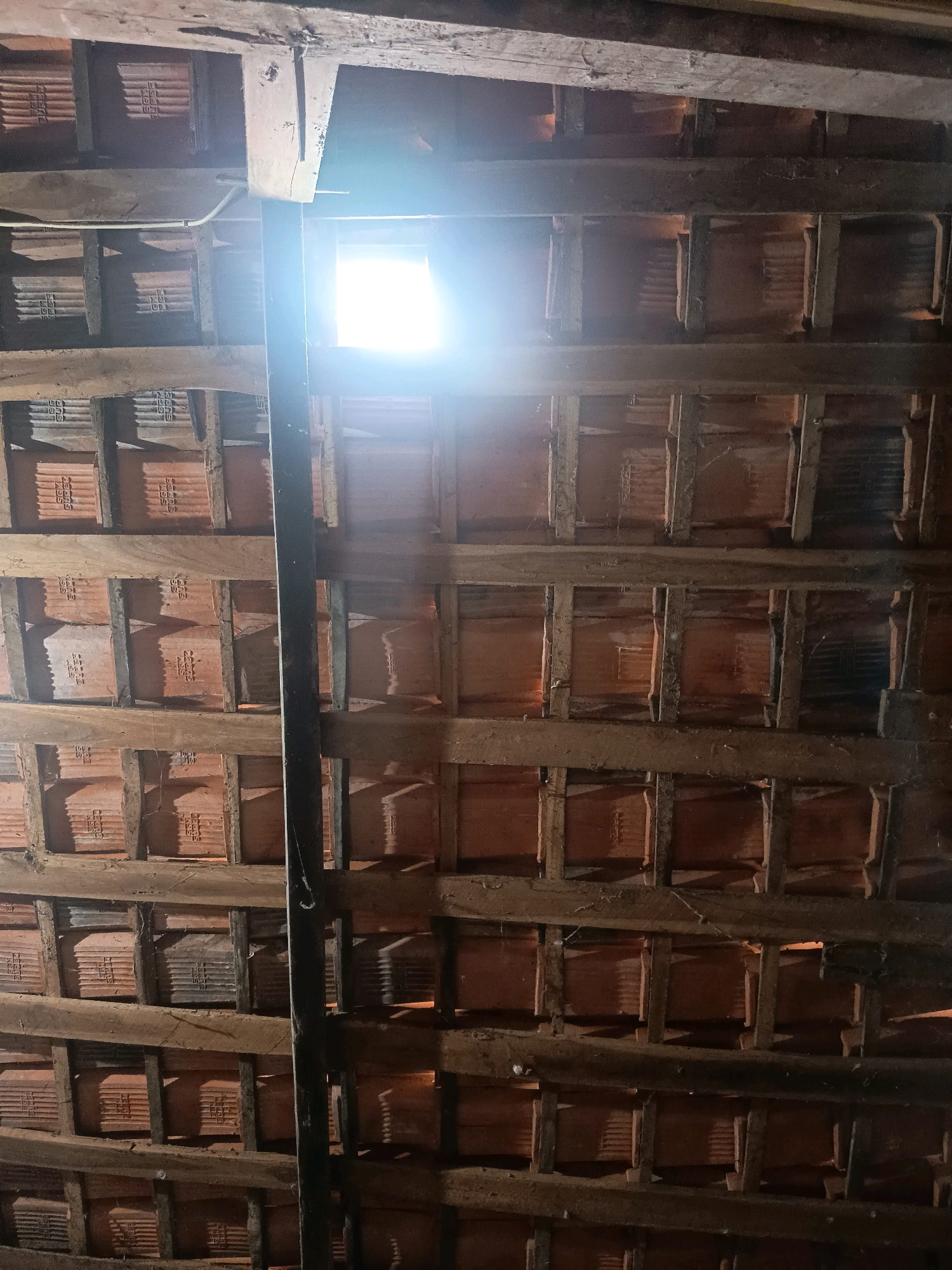 The image shows the underside of a tiled roof structure, likely in Sidorejo, Indonesia. It is a rustic, unfinished construction, featuring rough-hewn wooden beams supporting a layer of terracotta tiles. These tiles are laid in a grid pattern, with smaller wooden supports creating individual compartments for each tile. A prominent vertical black pipe or pole runs through the center of the frame, obscuring some of the view. A bright light source, possibly a window or opening, is visible near the top center of the image, casting a strong glare and illuminating the wooden beams and tiles. The overall impression is one of an old, possibly under-construction or disused attic space.