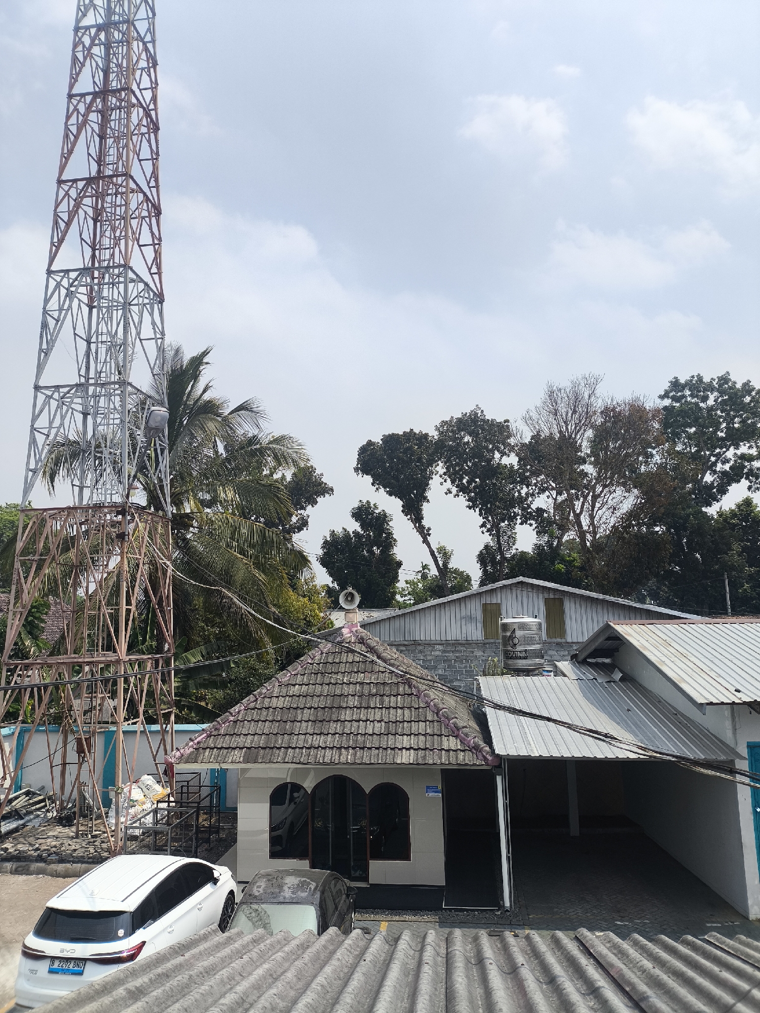 The image depicts an outdoor scene in Sukodono, Indonesia, dominated by a tall, metallic radio tower on the left. The tower has a lattice structure with some sections appearing reddish-brown and others silver. Below the tower, a row of buildings with tiled roofs is visible.

In the foreground, the roof of a building is covered with corrugated panels. Two cars are parked in the scene: a white electric vehicle on the left, with a license plate showing "B 2202 BND", and a dark-colored vehicle partially obscured behind it.

Further back, a small building with a distinctive pitched, tiled roof features arched windows and is painted white. Adjacent to this is a larger building with a metal roof. Trees, including palm trees, are scattered throughout the background, and the sky is partly cloudy. The overall environment suggests an urban or semi-urban setting.
