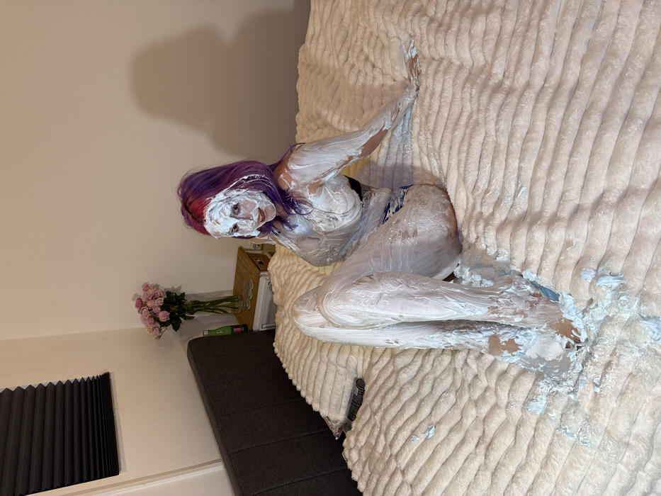 A person, covered in white cream, reclines on a plush, textured bed. Purple hair contrasts with the white substance on their face and body. The bed appears off-white, matching the cream. A nightstand to the side holds roses in a vase, with some objects on top. A dark, textured sofa is visible near a window with closed blinds in the background. The room's lighting is soft and even.