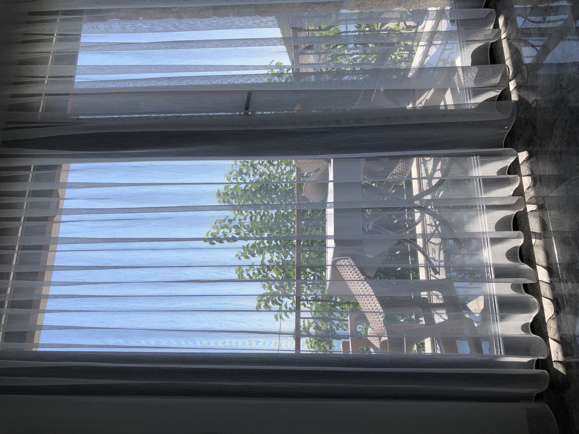 The image captures a view through sheer white curtains onto a balcony area. Through the semi-transparent fabric, one can discern a table and chairs on the balcony, suggesting an outdoor seating arrangement. Behind the balcony, a lush green foliage is visible, indicating the presence of trees or dense vegetation. Further in the background, a body of water, likely the sea, is apparent, with a hint of a hillside or landmass on the horizon. The lighting suggests it is daytime, with sunlight filtering through the curtains and casting shadows on the floor. The overall scene evokes a serene and pleasant atmosphere, possibly a view from a residence or a hotel room in a coastal location. The location is specified as Karaburun, Turkey, a town known for its coastal beauty.