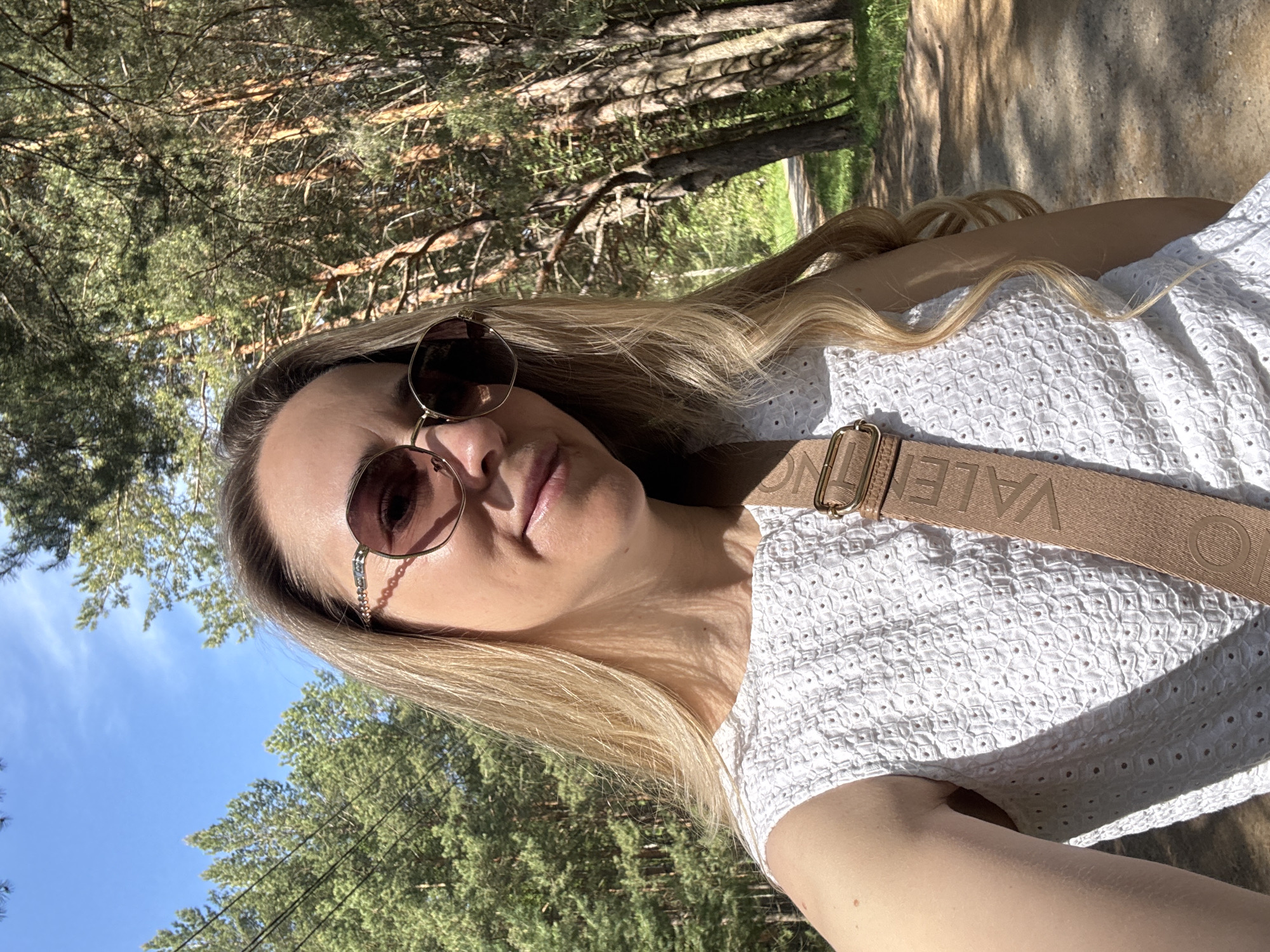 A blonde woman in sunglasses stands outdoors, likely in a wooded area, based on the trees visible in the background. She wears a white shirt with a textured pattern and a strap across her chest that has the word "VALENTINO" printed on it. The scene has clear lighting. Specific location information is unavailable.