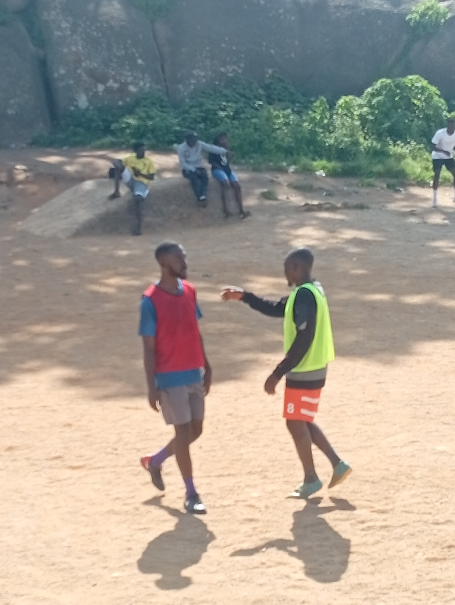In Jos, Nigeria, two young men are captured in a candid outdoor moment. The foreground is dominated by the two individuals, dressed in athletic attire. One man wears a red vest over a blue t-shirt and grey shorts. The other wears a bright green vest over a long-sleeved dark shirt and orange shorts with a white stripe and the number "8" on the side. Their poses suggest an interaction, possibly during a game or training session, with one extending his arm. The ground is a dusty, sandy surface. In the background, several other people are visible, some seated on a sandy incline, others standing, all casually dressed. Behind them, a large rock formation stands prominently, partially covered by lush green vegetation. The lighting indicates a bright, sunny day.