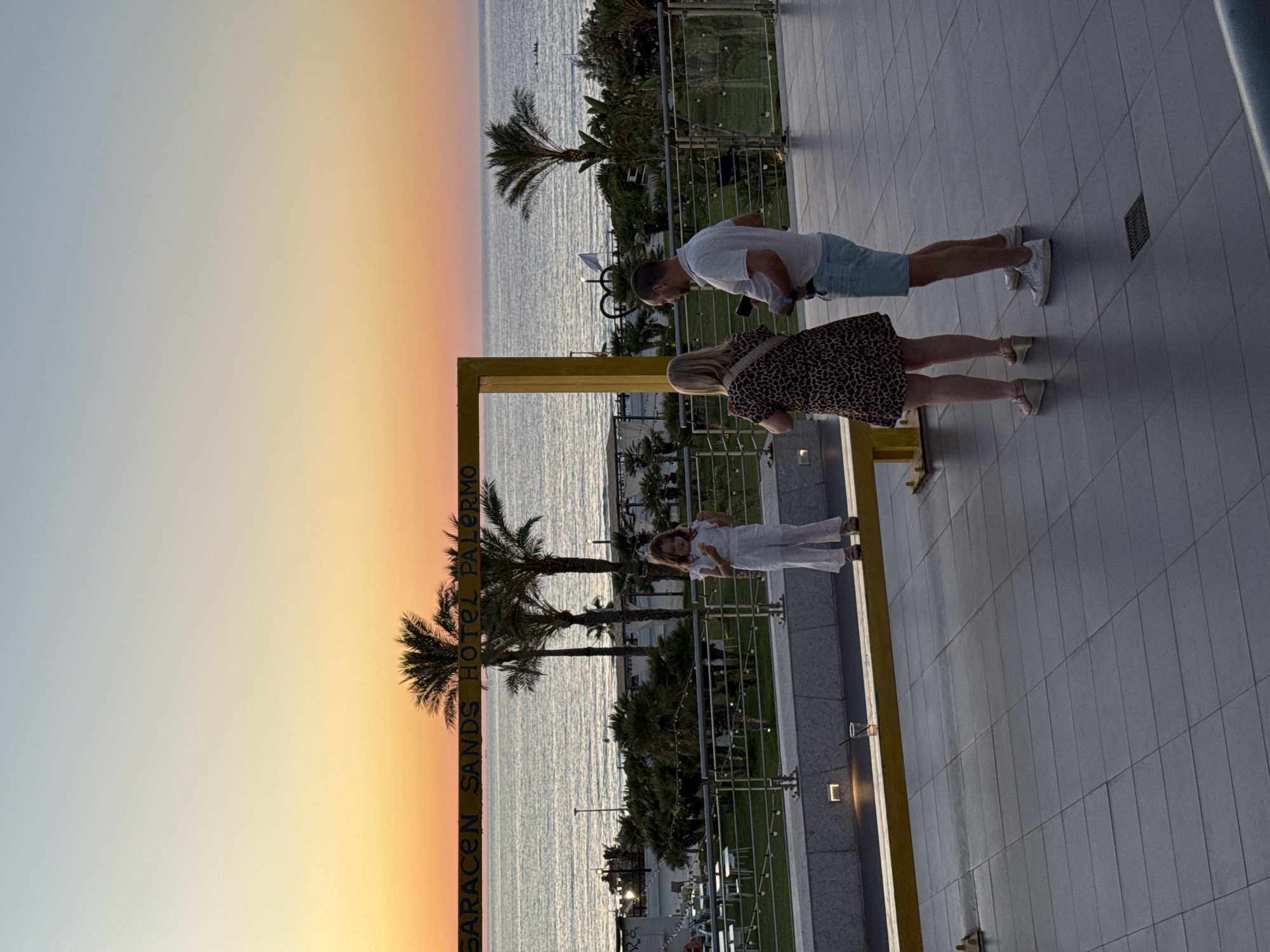 The image shows a sunset scene at Saracen Sands Hotel Palermo, located in Isola delle Femmine, Italy. A young girl in a white jumpsuit stands on a yellow beam, facing the ocean. Behind her, a couple is standing and looking out at the sea. The sky is a gradient of orange, yellow, and blue, indicative of a sunset. Palm trees and the Mediterranean Sea are visible in the background. The foreground shows a tiled patio and some outdoor seating. A glass with a straw and ice sits on the yellow beam near the girl.