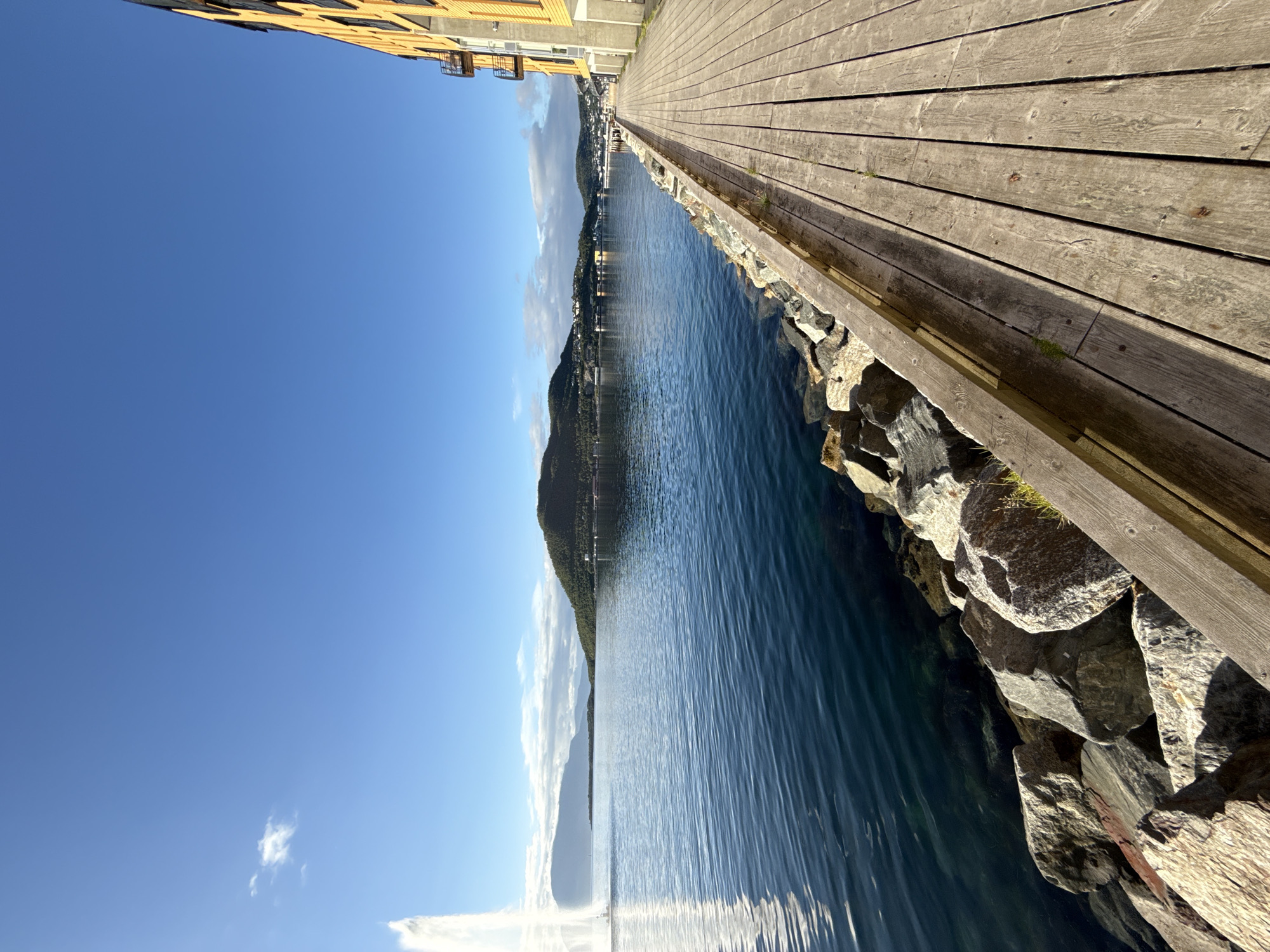 The image shows a waterside boardwalk next to a modern building in Harstad, Norway. The boardwalk is made of weathered wooden planks and runs along the right side of the frame, leading towards a body of water. The water is calm and deep blue, with ripples visible. A line of large rocks borders the water's edge. In the distance, a hill covered in green vegetation rises from the water, with buildings visible on its slopes and summit. A large fountain is visible in the water on the left side of the image, spraying a tall plume of water. The sky is a clear, bright blue with a few scattered white clouds. The building on the right is a multi-story structure with yellow siding and balconies, suggesting it is residential or commercial.