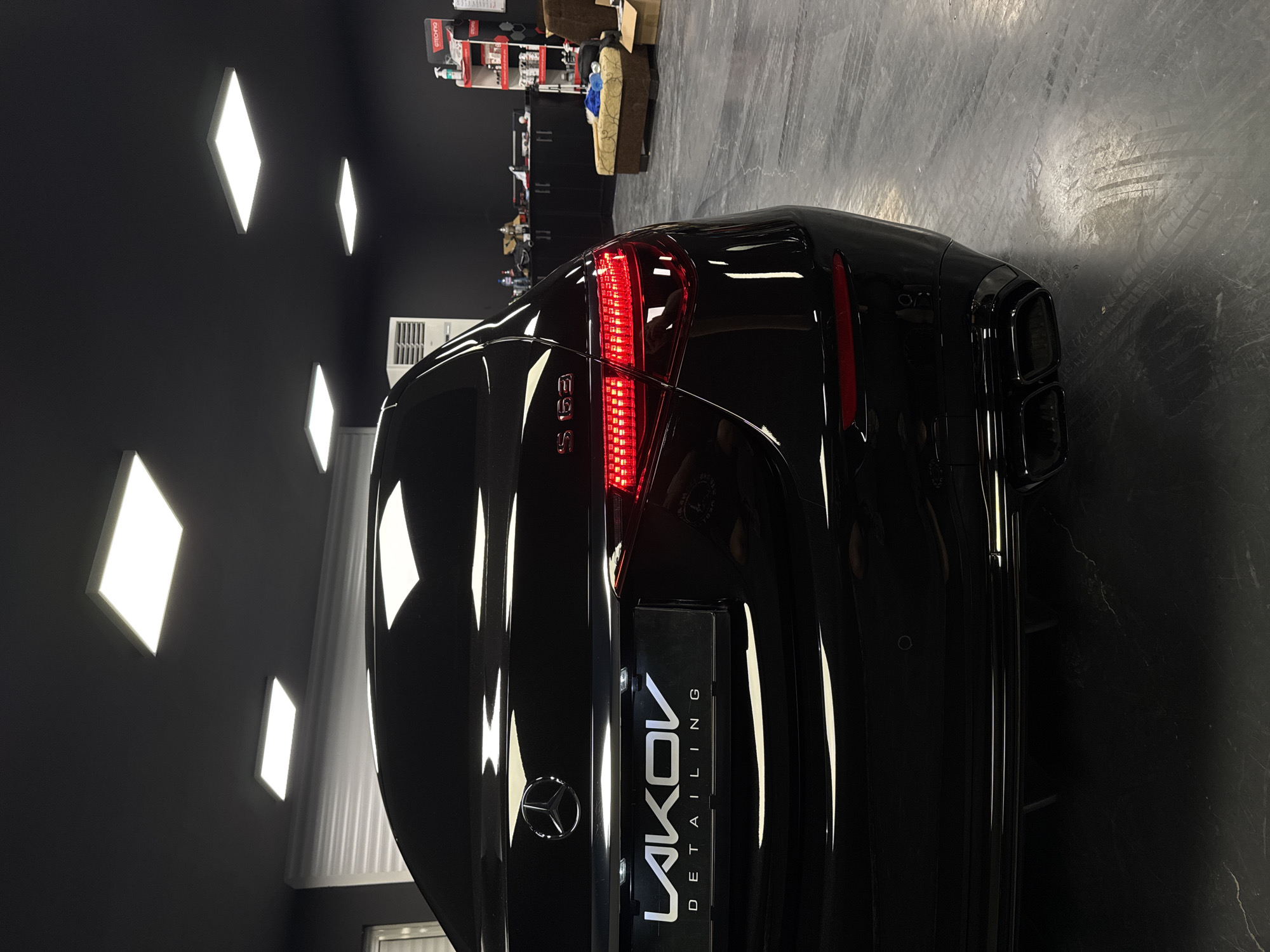 This is a low-angle, interior shot of a black Mercedes-Benz S63 AMG sedan parked in what appears to be a detailing or car storage facility. The car is positioned facing right, with its rear end prominently featured. The vehicle has its brake lights illuminated, displaying a red LED pattern. The "S63" badging is visible above the taillight on the right side. Below the Mercedes-Benz logo on the trunk is a custom license plate holder that reads "LAKOV DETAILING" in white stylized lettering. The car's twin exhaust tips are visible at the lower rear.
The environment is dimly lit, with a black ceiling featuring several illuminated square light panels. The walls are also black. To the right of the car, shelves filled with car care products are visible, along with a beige upholstered furniture piece and cardboard boxes. The floor is a dark, polished concrete with visible tire marks.
There are no people visible in the image. The overall impression is one of a well-maintained luxury vehicle in a professional, controlled environment. The specific location in the city of Ruse, Bulgaria, cannot be determined from the image alone.
