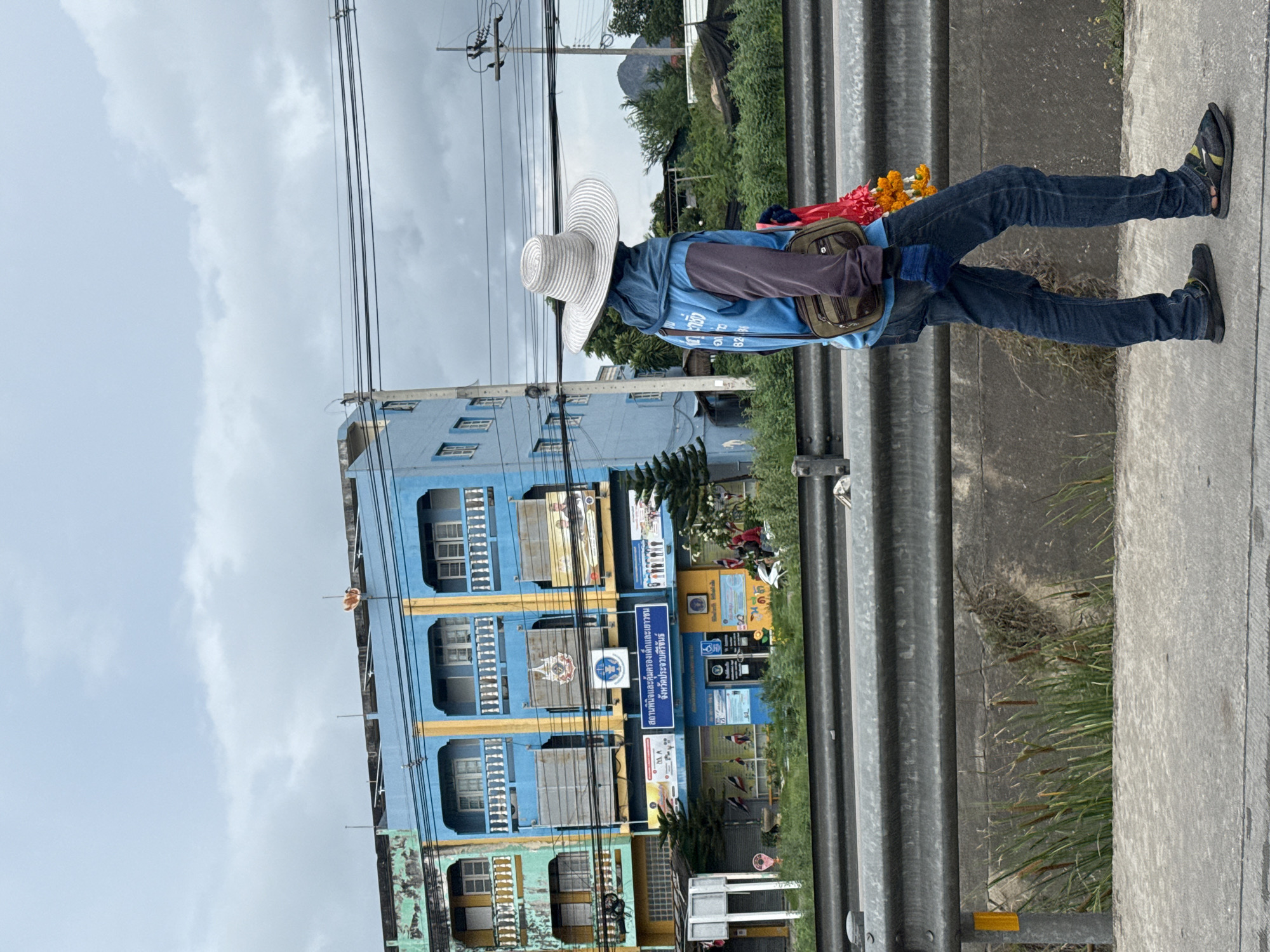 A person walks on a sidewalk beside a highway in Ban Nong Bua, Thailand, on a cloudy day. The individual, viewed from the side, wears a blue hooded jacket with dark sleeves, dark blue jeans, dark gloves, and a large, light-colored woven hat that obscures their face. A dark green crossbody bag is slung over their right shoulder, and a garland of red and yellow flowers hangs from their neck. In the background, a faded, multi-story building with blue and yellow facades stands, displaying various signs in Thai. Trees and power lines are visible between the building and the person. The foreground shows a concrete sidewalk, a metal guardrail, and dense green vegetation with cattails growing below the guardrail.