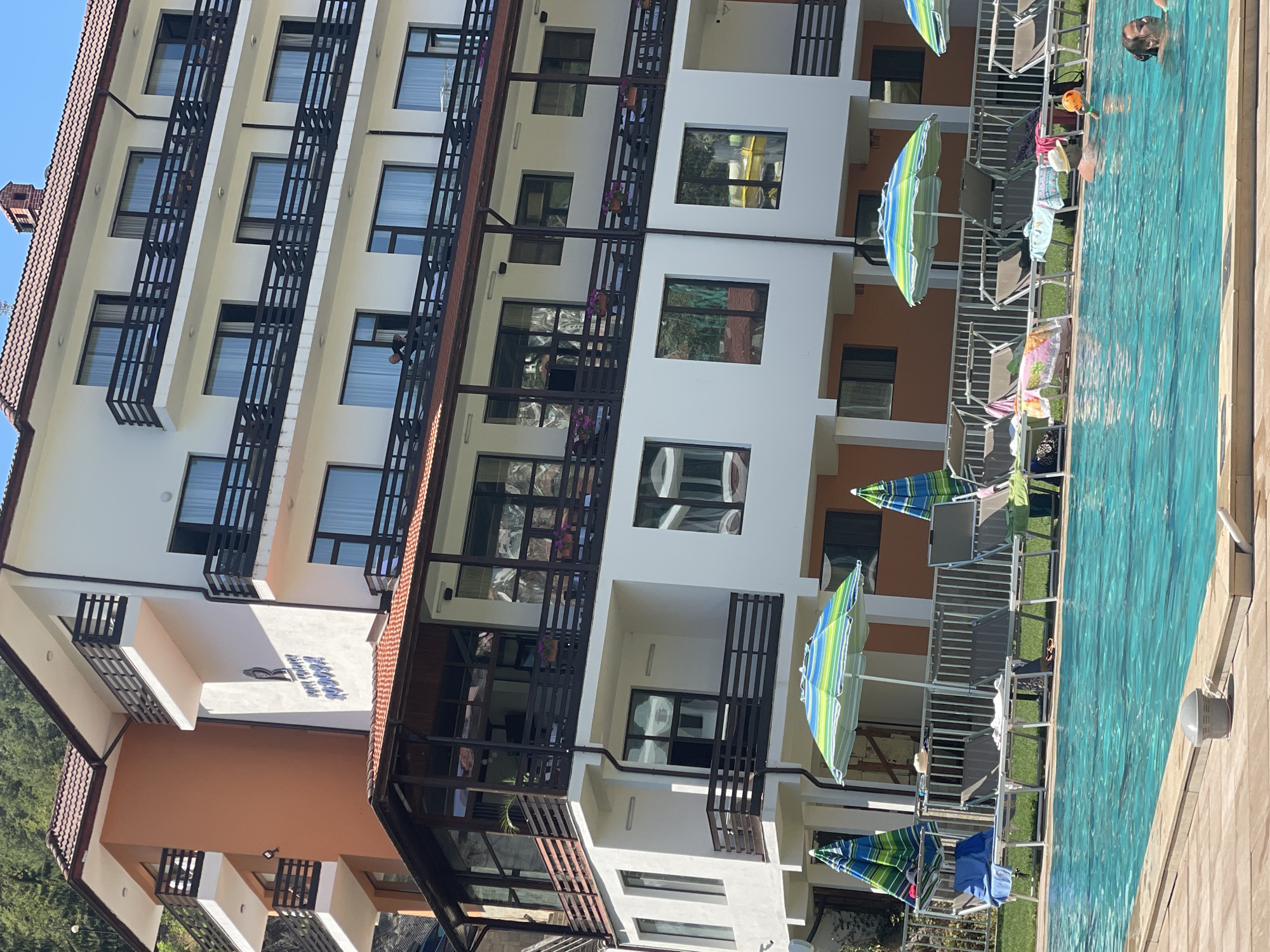 This image shows a hotel with a swimming pool in front of it. The hotel has multiple floors with balconies and windows. There are lounge chairs and umbrellas around the pool, and a few people are in the water. The hotel is situated in a natural setting, with trees and mountains visible in the background. The hotel has a sign that reads "ROPOТA". The city of Banite, Bulgaria, is mentioned as the location.