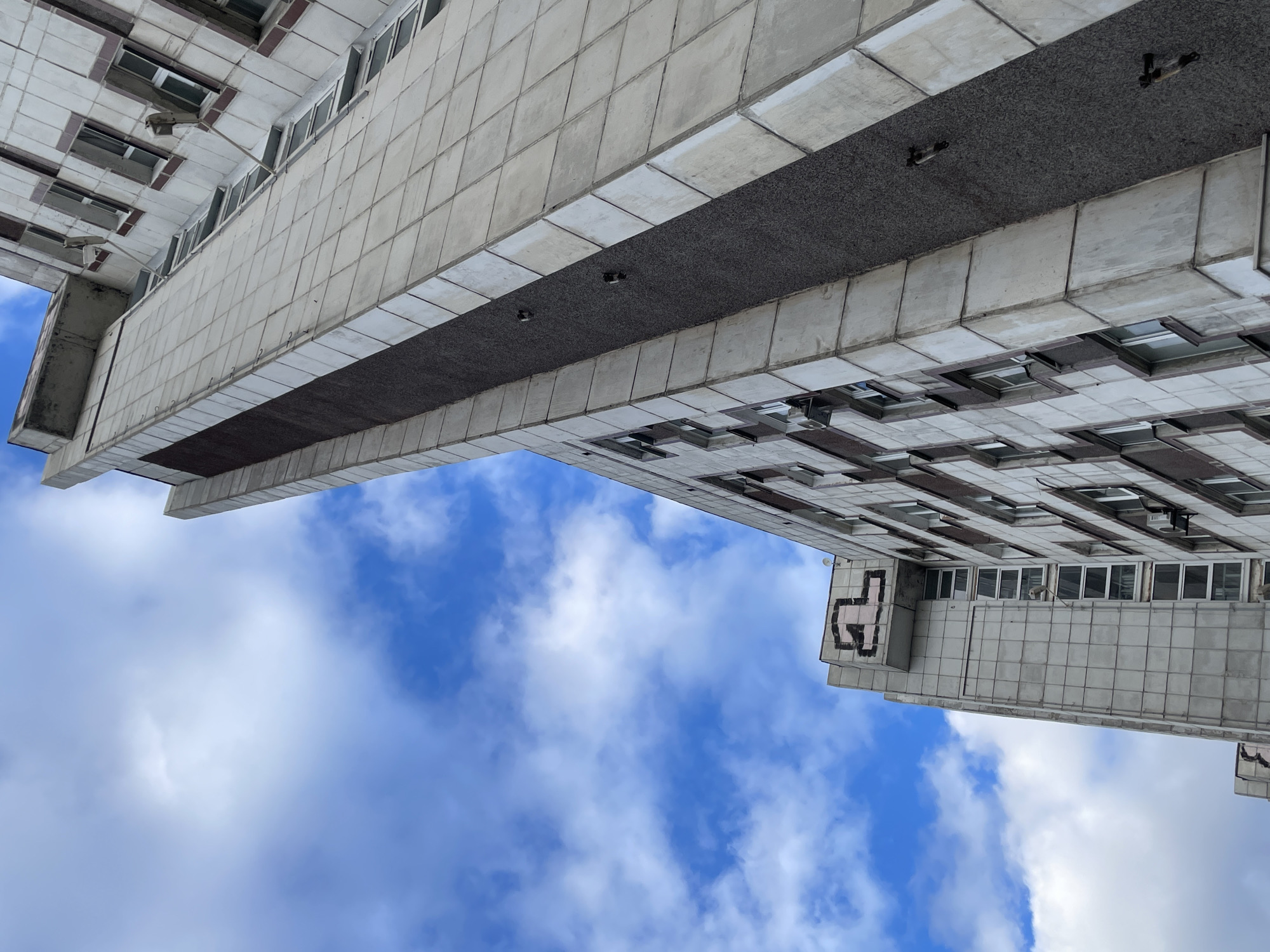 The image, taken from a low angle, showcases a section of a tall building against a backdrop of a blue sky dotted with white clouds. The building appears to be made of concrete, and its facade is characterized by a grid pattern of square or rectangular tiles. Windows are integrated into the grid pattern. A dark-colored band runs horizontally across the building, possibly indicating a change in the structural or design element. Graffiti is visible on one of the building sections. The overall impression is one of urban architecture juxtaposed against a natural sky. The location is Yekaterinburg, Russia.
