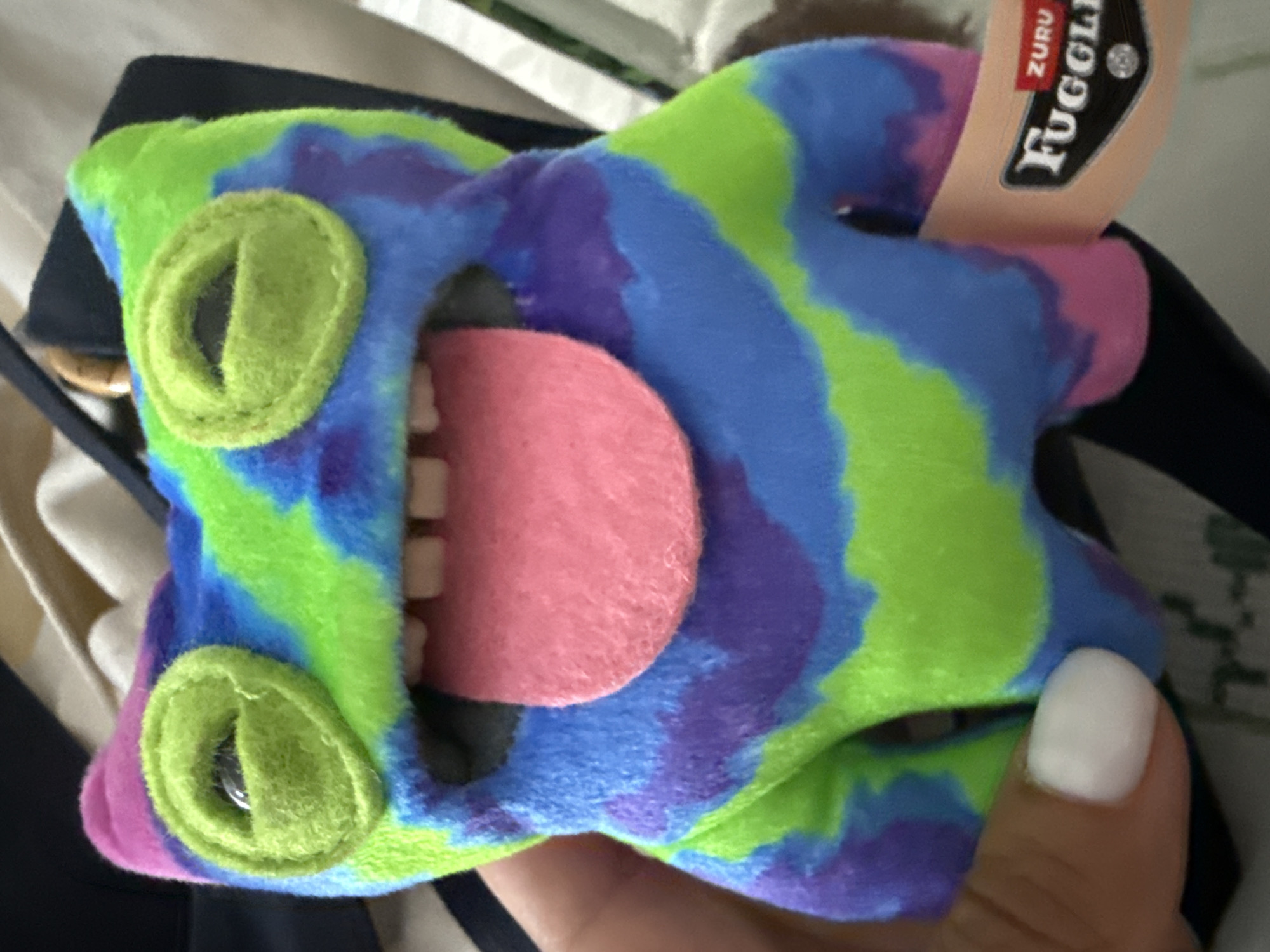 A close-up shot depicts a Zuru Fuggler toy held in someone's hand. The Fuggler is multi-colored in shades of green, blue, and purple, with two round, green button eyes. It has a wide open mouth exposing a pink tongue and white teeth. A Zuru Fuggler tag is attached to the toy. The background includes blurry images of clothing, including what appears to be a black jacket. The location is not identifiable from the image.