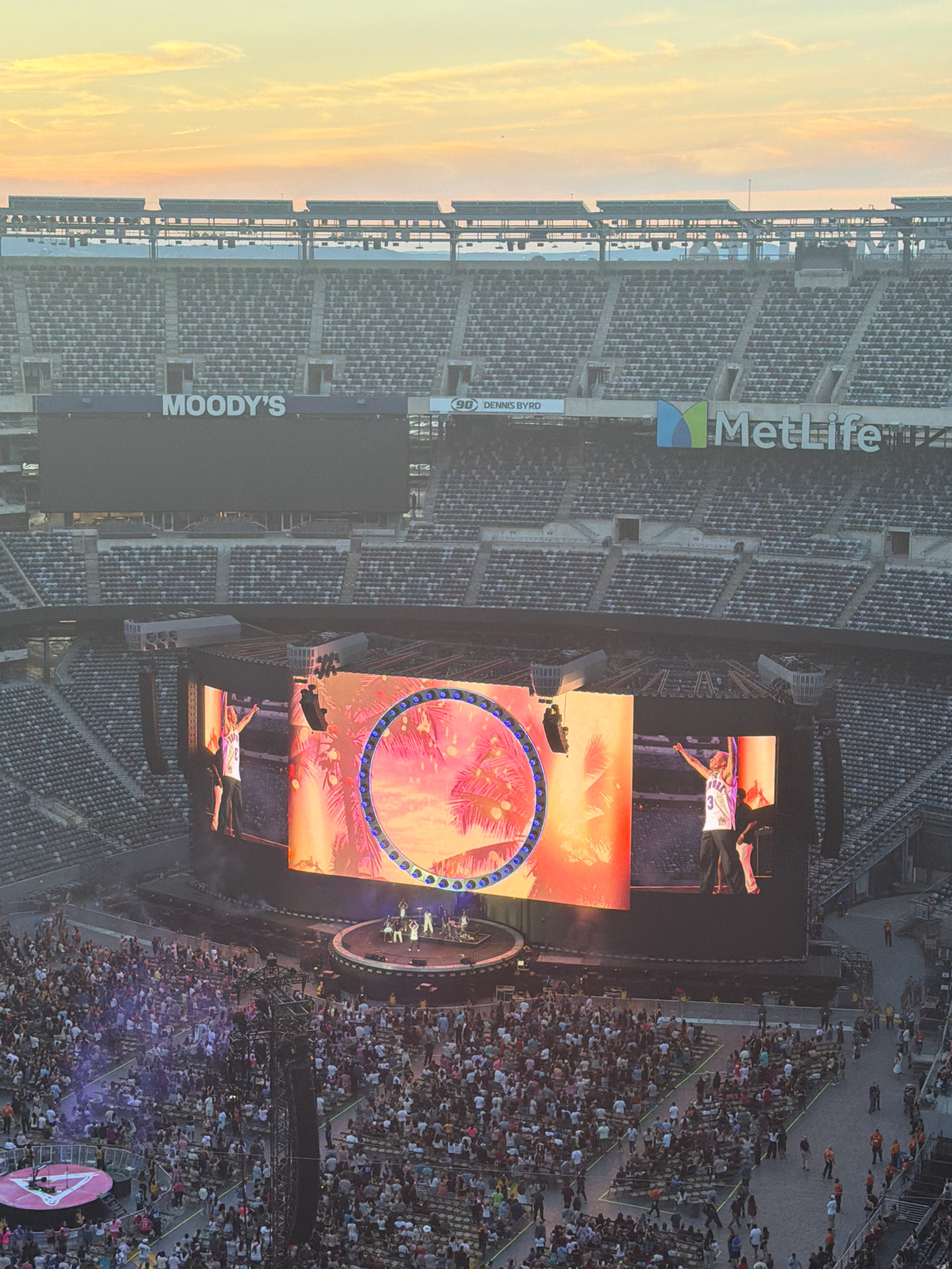 An aerial view captures a concert at MetLife Stadium in East Rutherford, New Jersey, United States of America, during sunset. The sky exhibits warm hues of orange, pink, and yellow, with scattered clouds. The stadium's seating is largely visible, with some sections populated by an audience. In the foreground, a large stage dominates the scene, featuring two massive LED screens displaying vibrant graphics of palm trees and tropical foliage within a circular frame. On the stage, performers are visible, including one individual wearing a white basketball jersey with the number "3" and the word "NEW YORK" across the chest, raising their arms. Below the stage, a dense crowd of concertgoers fills the stadium floor, illuminated by various lights. The stadium exterior displays prominent signage, including "MOODY'S" and "MetLife".