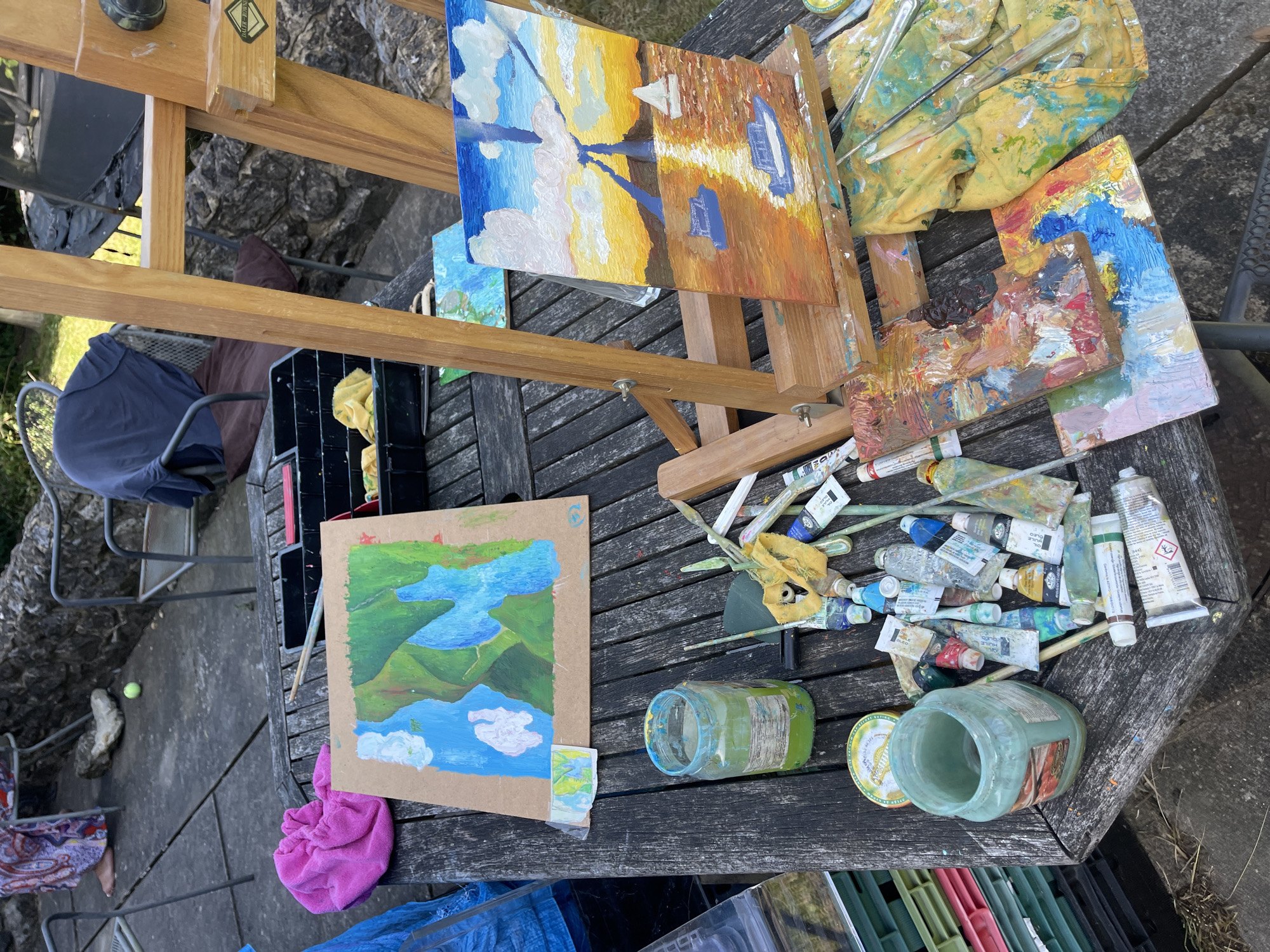 The image presents a high-angle view of an outdoor art-making space, likely a garden or patio. The scene appears to be set on a weathered wooden table, on which the artist's equipment is scattered. There is a wooden easel holding two paintings. One painting depicts a landscape with predominantly blue, white, and yellow colors, possibly representing the sky and reflections. The second landscape shows green hills and blue water. Tubes of paint, brushes, a paint-covered cloth, and jars (likely for water or solvents) are scattered nearby. A paint palette with colors, a tray with paint and a chair are present. The scene suggests an outdoor painting session in progress or recently concluded. The location is identified as Maidstone, United Kingdom.