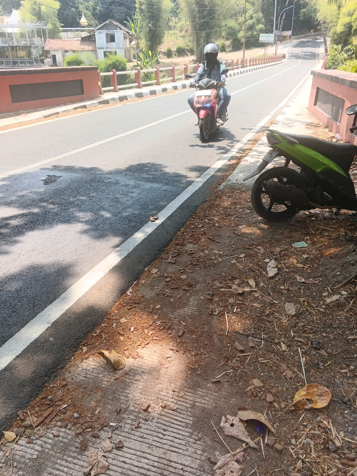 A lone person rides a red scooter on a paved road in a rural or suburban area of Semarang, Indonesia. The person is wearing a blue helmet and a dark jacket. The road has a white line marking the lane. On the right side of the road, there is a green scooter parked. The ground is covered with dirt, leaves, and debris. In the background, there are trees, buildings, and a bridge. The sun is shining, casting shadows on the road.