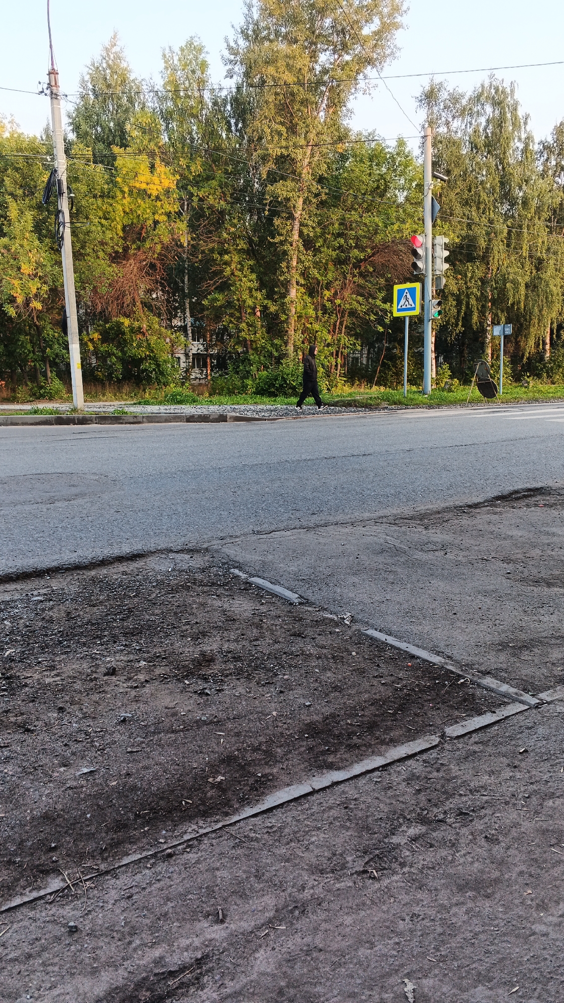 The image shows a street scene in Izhevsk, Russia, likely during the daytime, with a clear sky and sunlight illuminating the trees and road. In the foreground, there is a section of rough, dirt-covered ground adjacent to a paved road. The road itself is asphalt, with visible cracks and patches.
Further down the road, a pedestrian is walking across what appears to be a crosswalk. The pedestrian is dressed in dark clothing, including a hooded jacket, and is walking from left to right in the frame.
To the right of the pedestrian, there is a traffic light with a red light illuminated and a green light below it. Adjacent to the traffic light is a blue square sign with a white pedestrian symbol, indicating a crosswalk. Another sign, possibly a speed limit or directional sign, is visible further to the right.
The background features a dense line of trees with green and some yellowing foliage, suggesting it might be early autumn. Behind the trees, elements of a building are partially visible. A utility pole with electrical wires is present on the left side of the image. The overall atmosphere is urban, with a mix of natural and man-made elements.