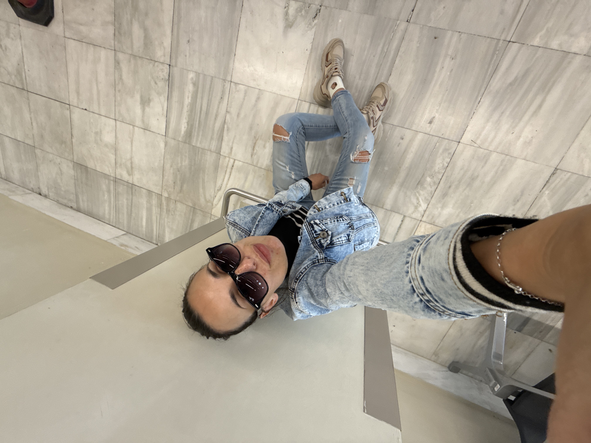 A woman is sitting on a chair, taking a selfie. She is wearing sunglasses, a denim jacket, ripped jeans, and sneakers. The background consists of tiled flooring and walls. The location is Kounoupidiana, Greece.
