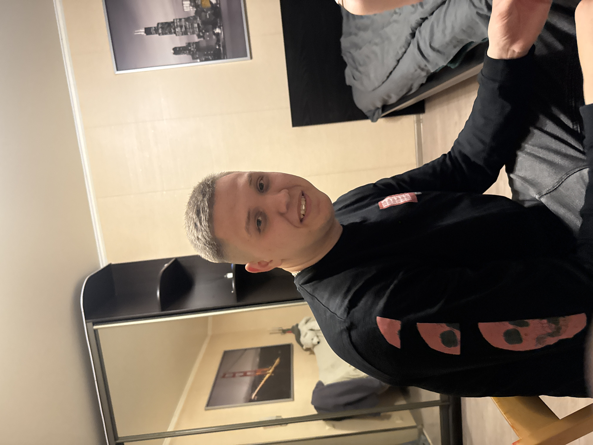 A man with short, graying hair is facing the camera and smiling slightly. He is wearing a black long-sleeved shirt with what appears to be a decorative pattern in reddish-pink on the sleeves.
Behind him is a room interior, including a sliding door wardrobe with a mirrored section. Reflected in the mirror is another room area containing a picture hanging on the wall. The primary room has a picture of a cityscape on the wall, and a portion of a bed with gray bedding is visible in the upper right corner.
The location information (Kazan, Russia) is not directly visible in the image, but is provided information.