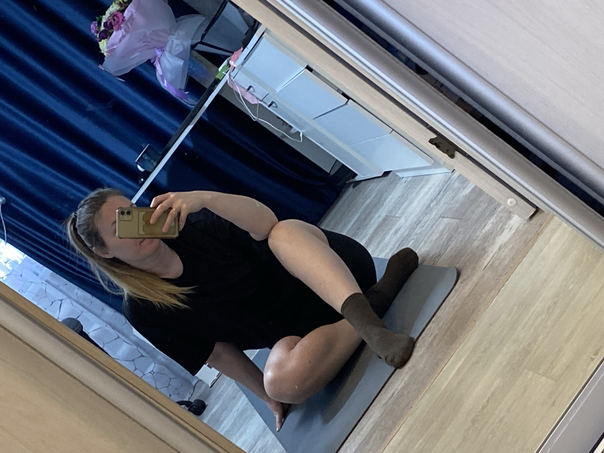 The image, taken in Noviy, Russia, captures a woman taking a selfie in a large mirror, depicting an indoor setting. The woman, with light-colored hair tied back, is partially obscured by the gold-colored smartphone she holds up to her face. She wears a black t-shirt and dark, possibly olive green, socks.
She is seated on a grey yoga mat on a light-colored wooden or laminate floor, with her legs bent in a relaxed position, suggesting she is either resting, stretching, or engaging in a light exercise.
The background, visible through the mirror's reflection, reveals several details of the room. Dark blue, pleated curtains hang on the left side. Above them, a bouquet of flowers wrapped in pink and white paper, containing purple and white blooms, is reflected. To the right of the curtains, a light-colored cabinet or shelf unit with multiple compartments is visible; a pink face mask with white elastic straps hangs from one of its shelves. The right side of the mirror's frame shows a metallic track, likely for a sliding door or wardrobe. A small, round white object rests on the floor near this track. In the bottom-left corner of the reflection, a wall with a light-colored, geometric, possibly tiled or textured pattern is seen, and a dark, rounded object lies on the floor nearby. The lighting appears to be natural, suggesting it is daytime. There is no visible text in the image.