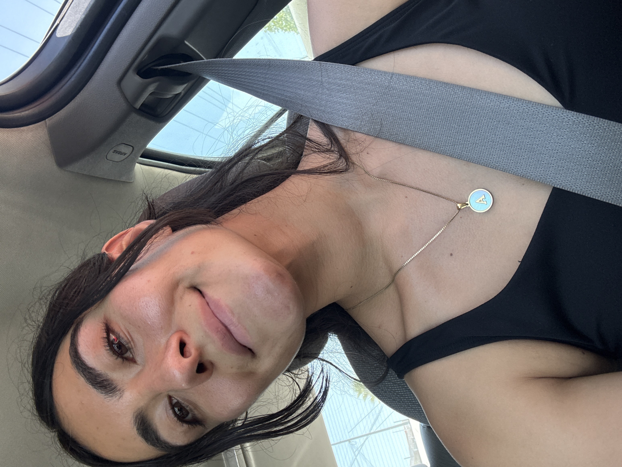 The image presents a close-up, angled shot of a person, likely a woman, inside a car in Puebla, Mexico. The subject has dark hair and dark eyes, and a fair complexion. Her face is turned slightly away from the camera, revealing a subtle, relaxed smile or smirk. She is wearing a black top or swimsuit and is secured by a gray seatbelt that crosses diagonally over her chest. A delicate gold-colored chain necklace with a small, circular, light blue pendant, featuring a white stylized "A" or similar emblem, rests on her chest.
The background shows elements of the car's interior, including a gray headliner and a plastic panel with the word "AIRBAG" embossed on it (visible upside down). Through the car's windows, a bright, clear blue sky and some blurred, light-colored structures are visible, indicating it is daytime. The activity suggests the person is either a passenger or driver, and the image appears to be a selfie or a candid moment captured within the vehicle.