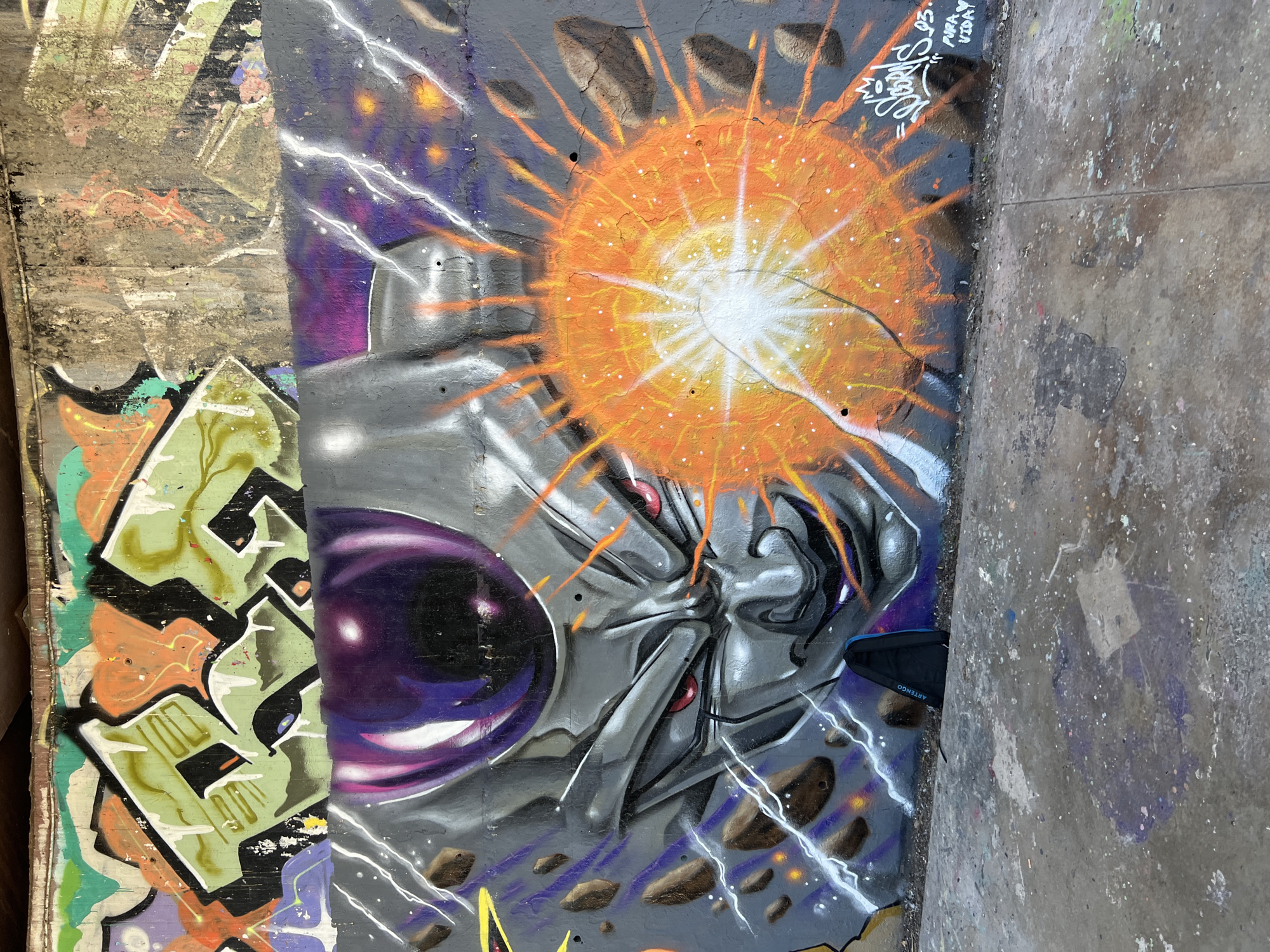 The image shows a graffiti mural depicting the character Frieza from the Dragon Ball Z anime. Frieza's head, with his signature purple head dome, red eyes, and a menacing expression, is prominently featured on the left side of the mural. He is shown preparing to launch a large, bright orange energy blast on the right side of the mural. The background is a dark, starry sky with streaks of purple and white lightning. Scattered rocks appear to be exploding around the energy blast, adding to the sense of destruction. The graffiti is painted on a concrete wall, which appears to be part of an urban environment. The ground in front of the wall is also concrete, covered in paint splatters and debris. A black backpack with blue accents rests against the wall at the bottom of the mural, near Frieza's neck. The mural is located in an outdoor, urban setting, possibly an underpass or a disused industrial area, judging by the textures of the wall and ground.
