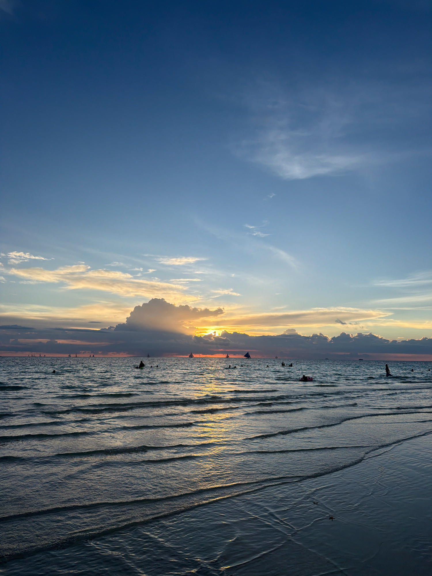 A wide-angle, vertical shot captures a sunset over the ocean at White Beach in Boracay, Malay, Philippines. The sky transitions from a deep blue at the top to lighter blues and oranges near the horizon. Wispy clouds are scattered across the sky, with a larger, darker cloud formation obscuring the setting sun. Beams of light emanate from behind this cloud, illuminating the sky with shades of yellow and orange.
The ocean is calm with gentle waves creating ripples on its surface, reflecting the sky's colors. Several sailboats with triangular sails are visible in the distance, dotting the horizon. Small groups of people are in the water, some swimming and others wading. One person is seen sitting in the water on a pink inflatable ring. Another person stands in the shallow water on the right side of the frame. The sandy beach is visible in the foreground, wet from the receding tide, with intricate patterns formed by the water's edge. The overall scene depicts a serene evening at a popular beach destination.