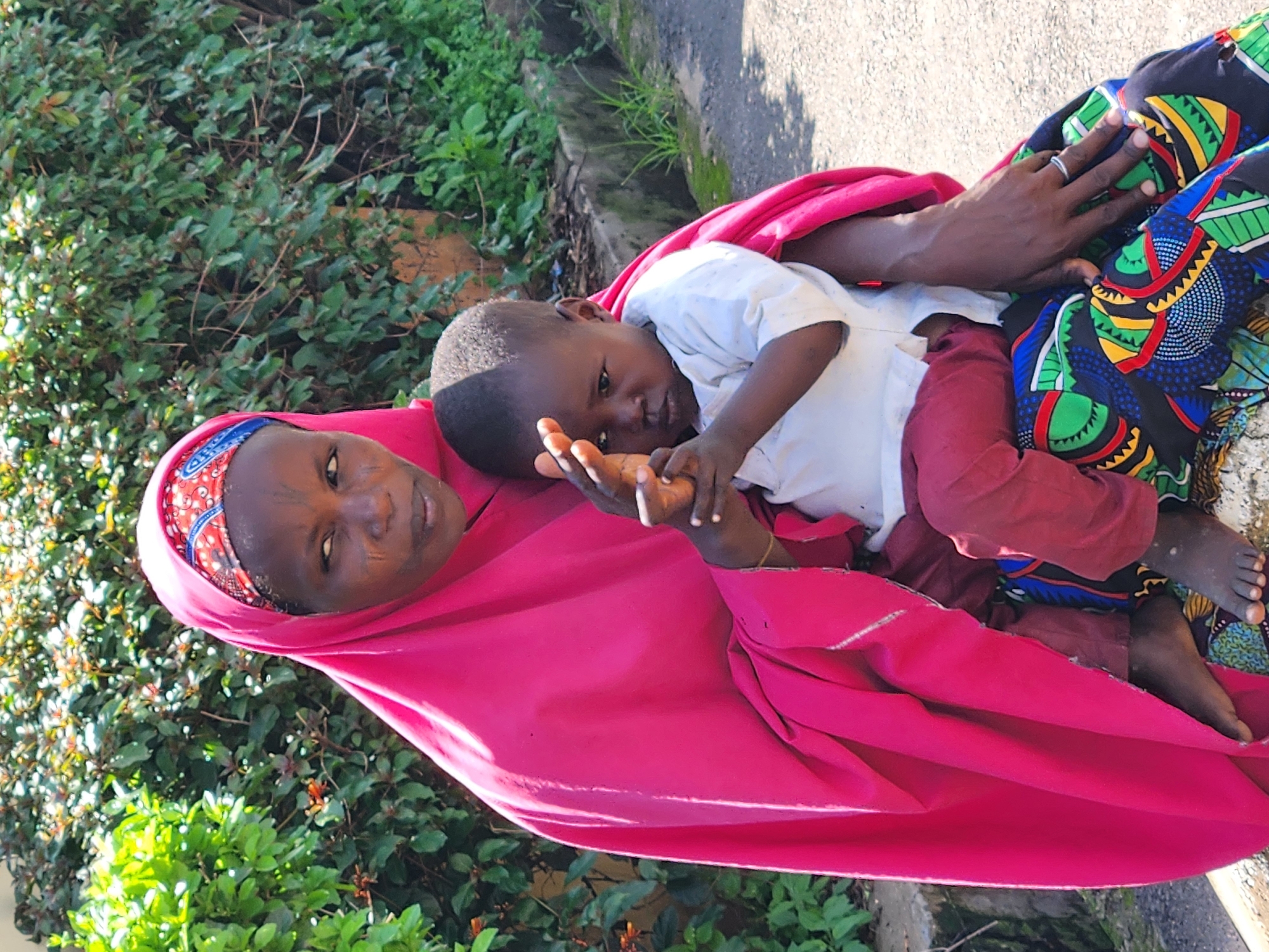 An adult woman wearing a bright pink hijab and an patterned skirt is seated outdoors with a young child on her lap. The woman has facial markings and is looking to her left. The child is wearing a white shirt and red pants and is looking towards the viewer with their right hand raised. The background consists of lush green foliage and a paved surface. The scene is well-lit by sunlight.