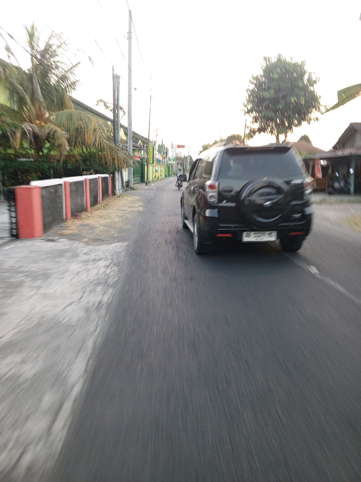 The image depicts a street scene in Bantul, Indonesia, likely in the afternoon given the lighting. A black SUV with a spare tire mounted on the back is driving away from the camera on an asphalt road. The SUV has a license plate that appears to read "B 1225 MG". To the left of the road, there is a concrete sidewalk with a low wall or barrier constructed from concrete pillars painted red and dark gray, interspersed with dark gray panels. Behind this wall, palm trees and other foliage are visible. Further down the street, more buildings and some power lines can be seen. A motorcycle is also visible in the distance, ahead of the SUV. The sky is bright and appears overcast or hazy. The image has motion blur, indicating it was taken from a moving vehicle.