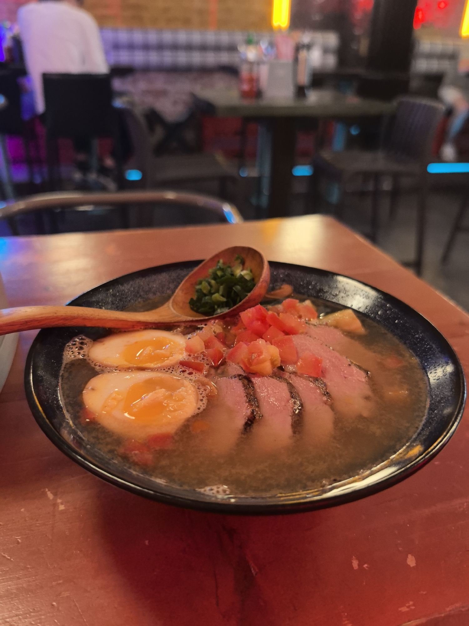 The image shows a close-up of a bowl of ramen on a red table. The ramen contains sliced meat, two halved soft-boiled eggs with runny yolks, chopped tomatoes, and green onions. A wooden spoon rests on the edge of the bowl, holding a small portion of green onions. In the blurred background, a restaurant setting is visible with tables, chairs, and patrons. The overall lighting suggests an indoor environment, possibly during the evening. There is no indication of the city of Perm, Russia, in the image itself.