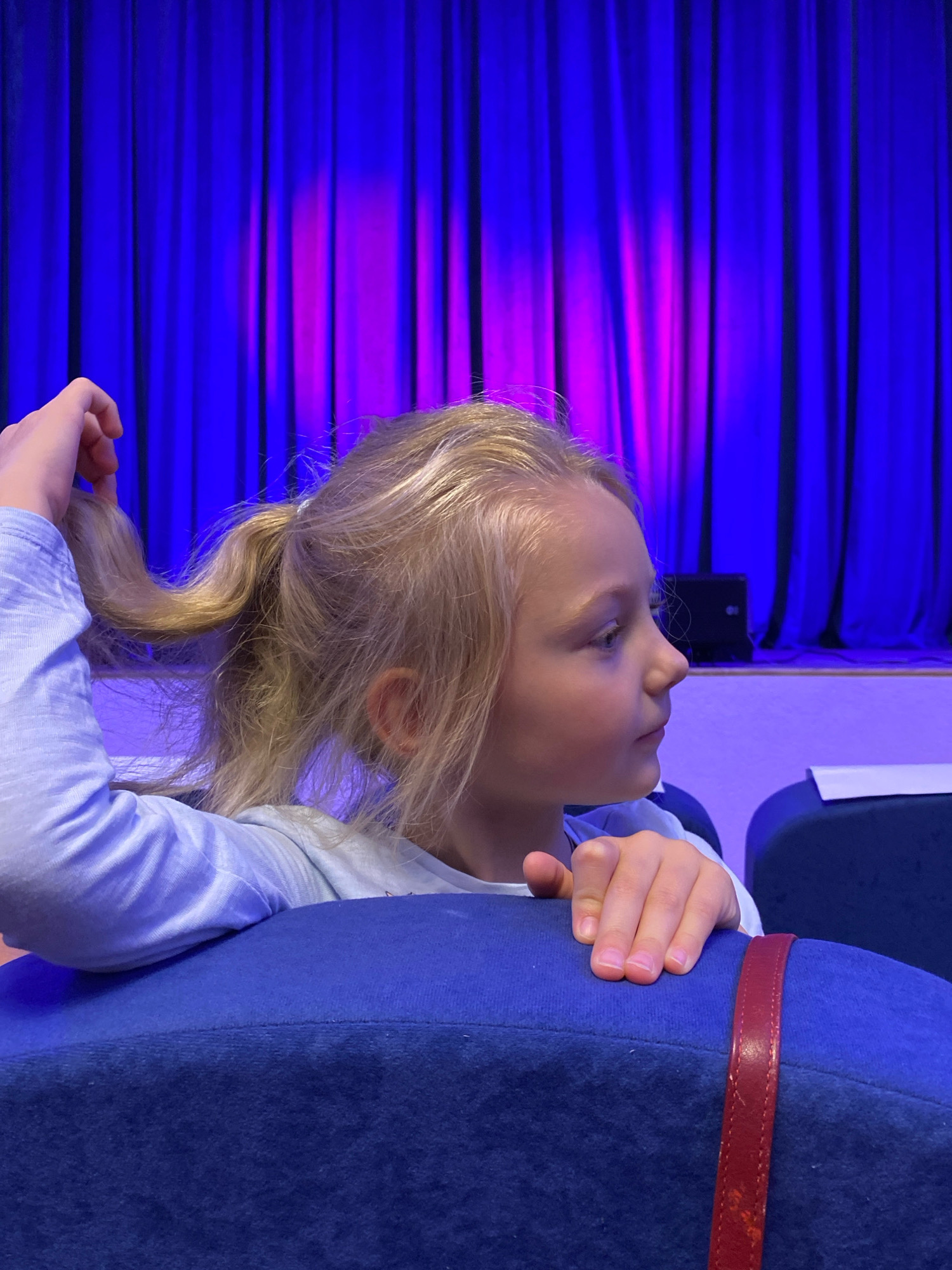A young girl with blonde hair pulled into a ponytail, wearing a light blue shirt, is seen from the side in a theater. She is sitting on a blue seat, with her arm resting on the back of it and her hand near her face, holding a lock of her hair. In the background, a stage is visible with blue and pink curtains and a speaker. There is no information in the image to suggest the location is in Ivanteyevka, Russia.
