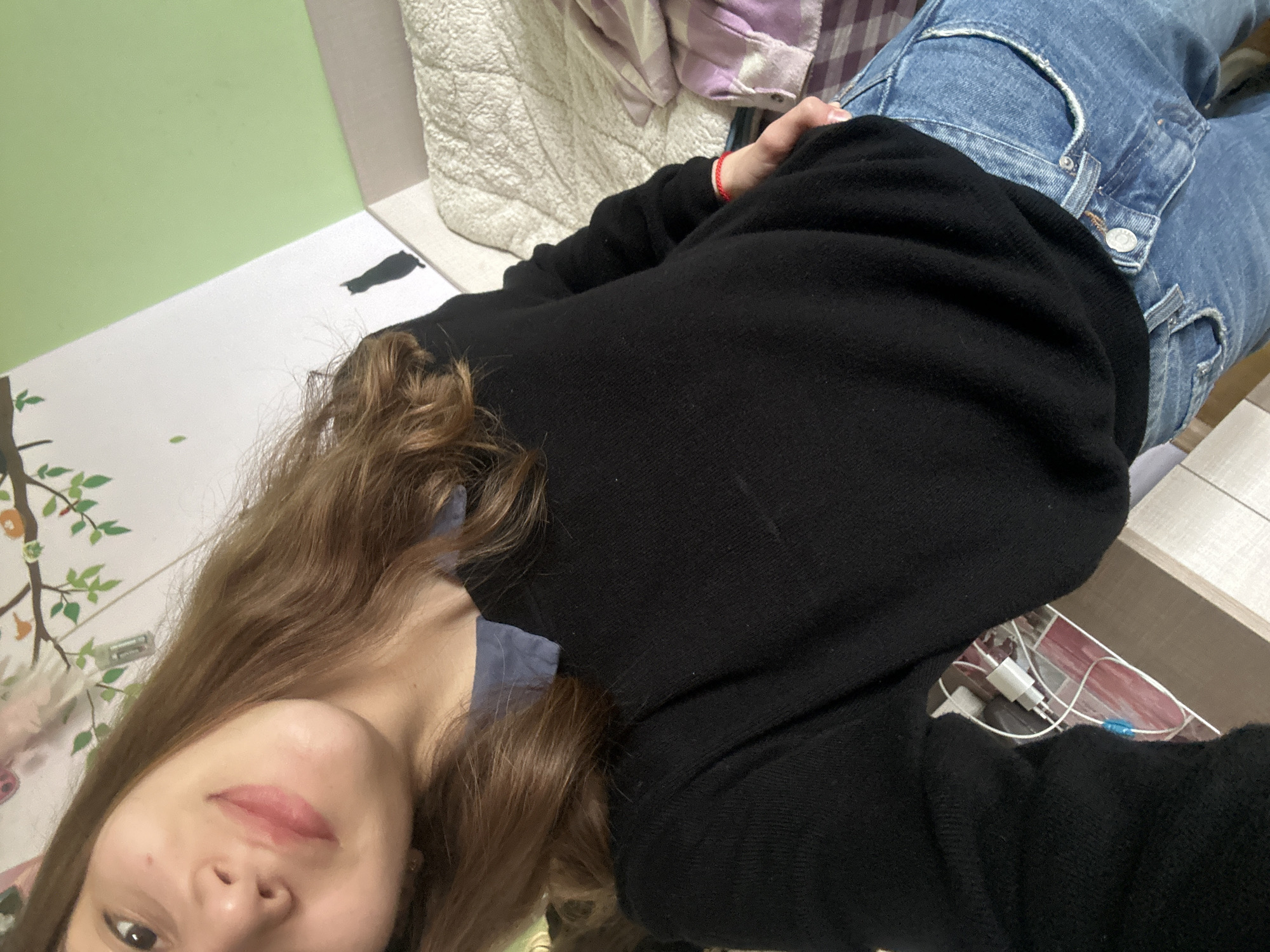 A young woman lies at an angle on a bed or floor in an interior room. She is wearing blue jeans and a black sweater. Her long, brown hair falls around her face and shoulders. Part of the room's wallpaper includes a tree design. A cell phone charger is visible near her. It is impossible to determine the location in the city of Ruse, Bulgaria from this image alone.