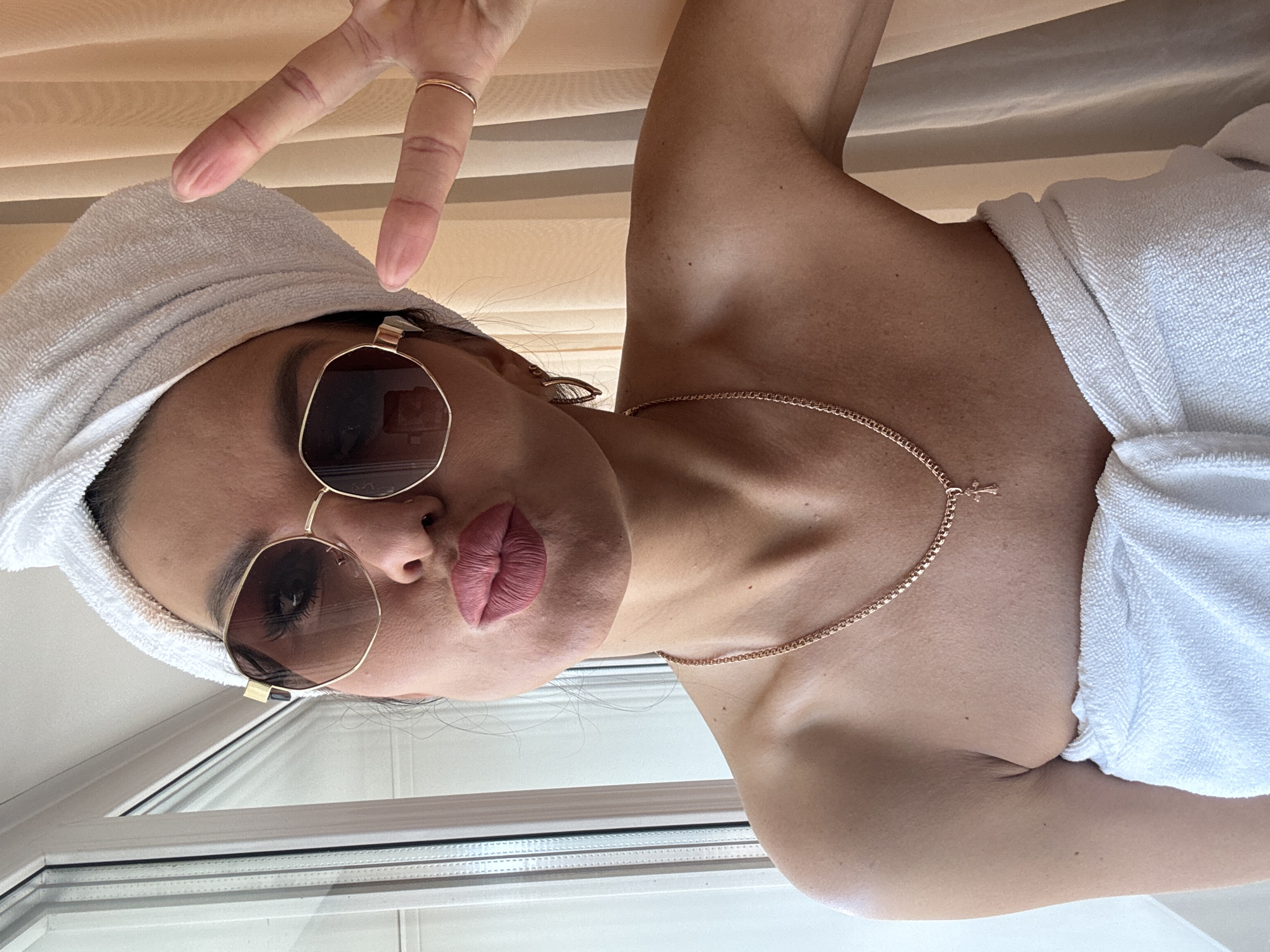 A woman poses indoors, presumably near a window, in Perm, Russia. She is wearing a white towel wrapped around her head and another one draped around her shoulders. She is wearing rose-tinted sunglasses and a gold chain necklace. She is making a peace sign with her left hand. The background includes window blinds and a window frame.