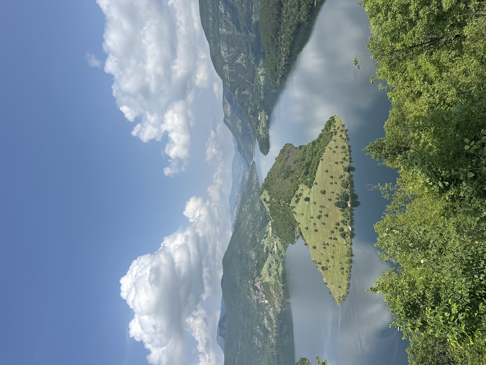This media file presents a scenic panoramic view of a large body of water, likely a reservoir or lake, nestled amidst forested mountains and hills under a bright blue sky. The image appears to be taken from an elevated viewpoint, possibly looking down into a valley or gorge in Ribartsi, Bulgaria.
The sky, occupying a significant portion of the upper left, is a vibrant blue dotted with large, fluffy white cumulus clouds. These clouds cast soft reflections upon the calm surface of the water below. The main body of water stretches across the middle and lower right of the frame, its surface largely still with subtle ripples that suggest a gentle breeze.
Lush green mountains and hills, densely covered with trees, rise dramatically from the water's edge on both sides, receding into the hazy distance where further mountain ranges can be faintly seen. A prominent, elongated landmass—possibly a peninsula or a large island—covered in bright green grass with scattered small trees, juts out into the water from the left side, dividing the water body.
Along the distant shores, small clusters of buildings or villages can be discerned amidst the greenery, indicating human presence in this otherwise natural landscape. In the immediate foreground on the right, vibrant green foliage from trees partially frames the scene, suggesting the photographer was positioned behind or among trees.
The time of day appears to be bright daylight, likely late morning or early afternoon, given the clear illumination and the nature of the clouds. The weather is clear and sunny, contributing to the serene and picturesque atmosphere. There is no visible text in the image. The overall impression is one of natural beauty and tranquility in a mountainous, waterside setting.