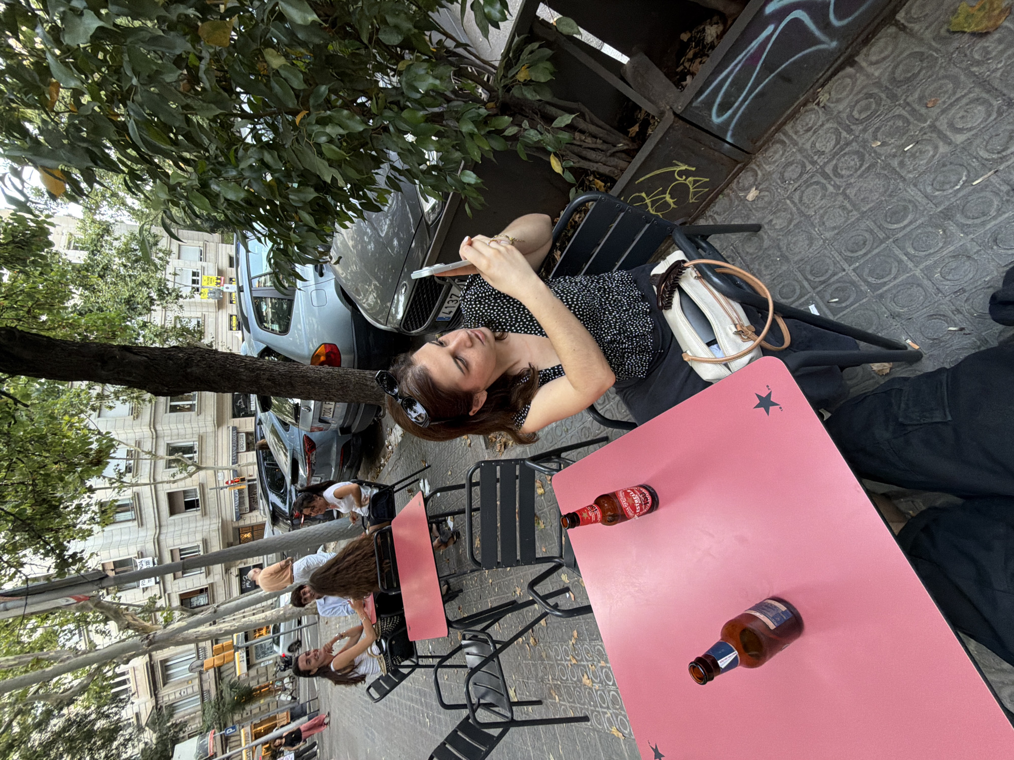 This media file shows a street scene in Barcelona, Spain, likely in the afternoon or early evening. Several people are seated at tables outside a cafe or restaurant.
In the foreground, a pink table has two bottles of Estrella Damm beer on it. A person, facing away from the camera, is sitting at a black chair with their legs crossed. Their right hand is holding a smartphone. They are wearing a black and white polka dot top and dark pants. A light-colored purse is also visible on the chair.
In the background, other people are seated at tables. One person is wearing a white t-shirt and looking towards the right of the frame. Another person in a light orange shirt is visible. The scene includes trees with green leaves and a street with parked cars and buildings lining the sidewalk. The ground is paved with patterned tiles. Graffiti is visible on a metal structure to the left of the main subject. The overall atmosphere appears casual and relaxed.