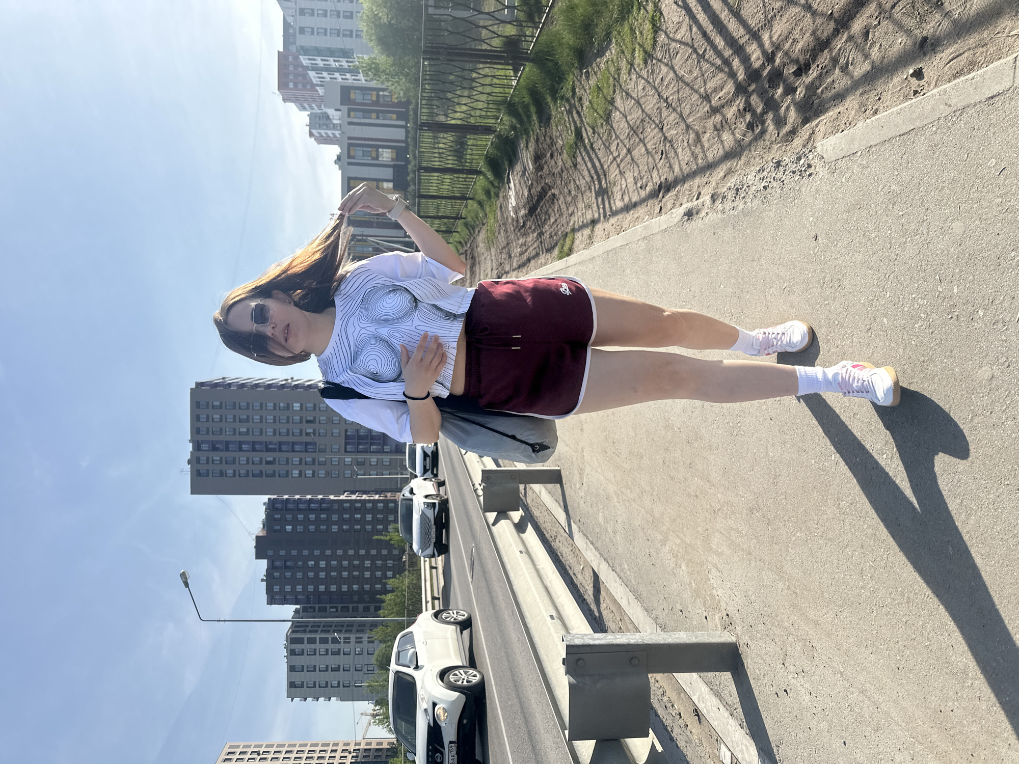 A young woman stands on a city sidewalk in Perm, Russia. She wears sunglasses, a graphic tee, maroon athletic shorts, white socks, and white sneakers. Her hair is long and brown. She is standing on a sidewalk next to a road, with cars parked along the street. In the background are tall apartment buildings under a bright sky.