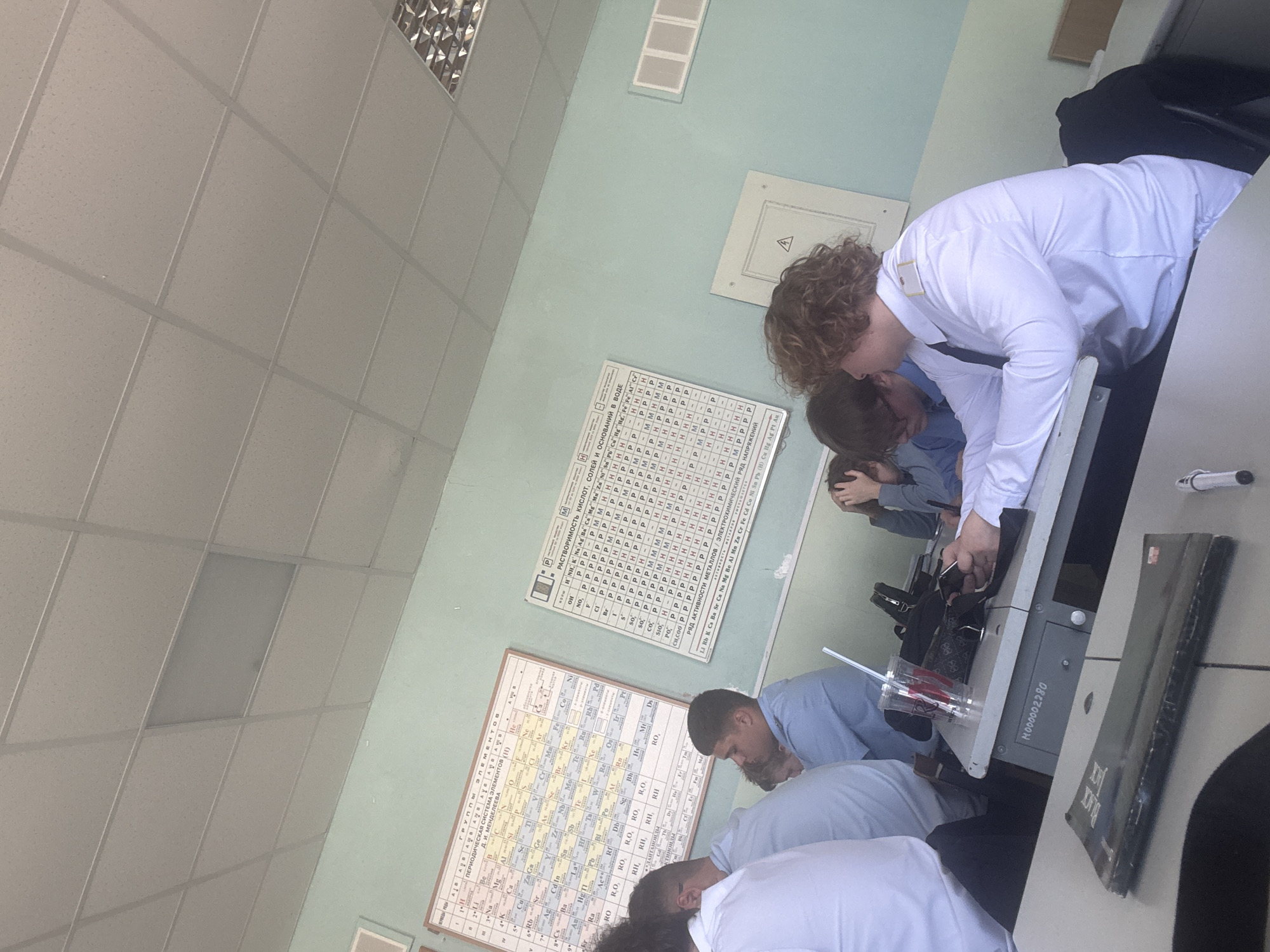 The image captures an indoor scene, likely a science classroom or laboratory, with several students engaged in an activity at desks. The room features light blue-green walls and a white ceiling with square acoustic tiles and fluorescent lighting fixtures.
In the foreground, a student with curly brown hair, wearing a white long-sleeved shirt, leans over a desk, seemingly looking into or operating a piece of lab equipment, possibly a microscope. Behind him, other students are also bent over, focusing on their respective stations. One student is seen in a light blue shirt, and another further back has their hands near their head.
Notable objects on the desks include a clear plastic cup with a red straw, a white marker or pen, and a black book partially visible with "BLACK BOX" on its spine. One desk has a label "N000002280".
Two large educational posters hang on the wall. The upper poster, titled "РАСТВОРИМОСТЬ КИСЛОТ, СОЛЕЙ И ОСНОВАНИЙ В ВОДЕ" (Solubility of Acids, Salts and Bases in Water), displays a grid indicating the solubility of various chemical compounds. Below it, a poster titled "ГРУППЫ ЭЛЕМЕНТОВ / ПЕРИОДИЧЕСКАЯ СИСТЕМА ЭЛЕМЕНТОВ (Н)" shows a periodic table and related chemical information, including "РЯД АКТИВНОСТИ МЕТАЛЛОВ" (Activity Series of Metals).
The lighting suggests daytime, and the overall atmosphere is one of focused learning or experimentation.