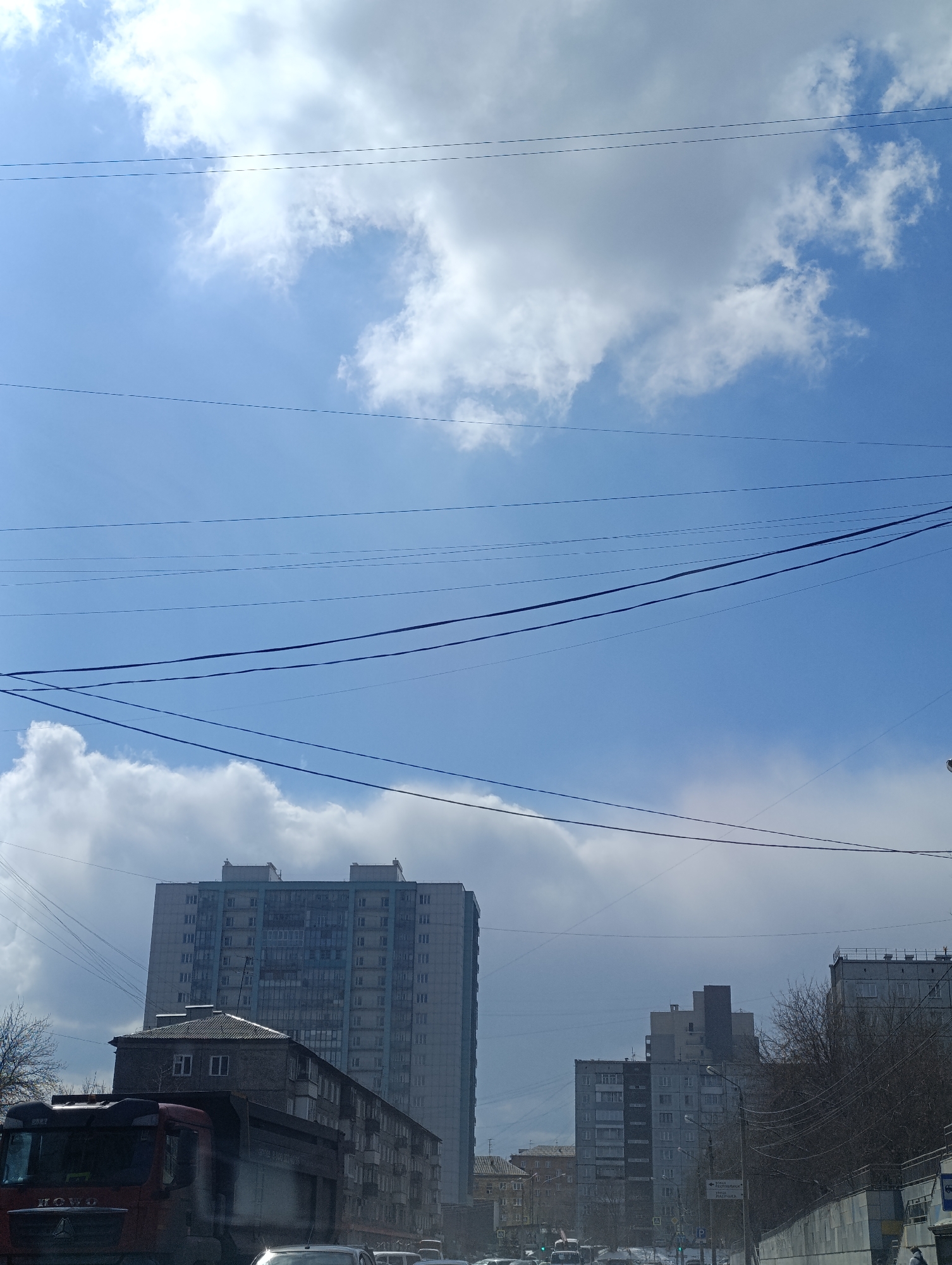 This image captures a daytime urban street scene in Krasnoyarsk, Russia, under a partly cloudy sky. The perspective appears to be from inside a vehicle, possibly moving, indicated by reflections and the angle of view.
The upper half of the image is dominated by a bright blue sky with large, white cumulus clouds. Numerous black electrical and utility wires crisscross the sky, creating a grid-like pattern.
Below the sky, the landscape features a mix of architectural styles. On the left, a large, modern-looking multi-story apartment building with many windows stands tall, appearing somewhat faded. In front of it, an older, smaller, darker-colored residential building with a sloped roof and brick facade is visible, partially obscured by a large red truck in the foreground. This truck, visible from its front and side, has "HOWO" clearly written on its grille, indicating it's a Chinese-manufactured heavy vehicle.
Further down the street to the right, several other multi-story apartment buildings are visible in the middle and far distance, mostly in shades of gray and beige, characteristic of Soviet-era construction. Bare tree branches suggest a late winter or early spring season.
Traffic appears to be present on the road, with glimpses of other cars and vehicles. A green road sign in the distance displays Cyrillic text, reading "ПЕР. АСТАФЬЕВА" (Per. Astafyeva - Astafyev Lane) and "НАСЫПНАЯ" (Nasypnaya - Embankment). Another sign shows a bus stop symbol and what looks like parking information. The overall impression is that of a bustling urban environment on a clear, bright day.