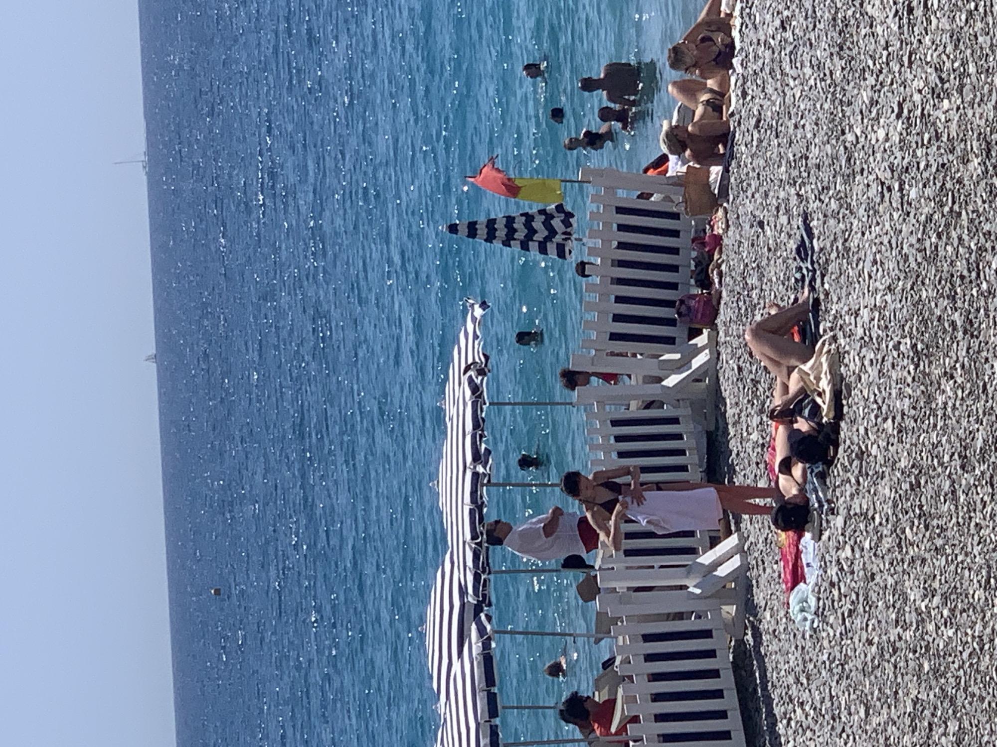 This image captures a vibrant beach scene on the pebbly shore of Nice, France. In the foreground, several individuals are sunbathing on towels spread across the stones. One woman in a black bikini lies on her front, while another next to her reclines on her side. Further up the beach, more people are visible, some seated and others reclining.
The middle ground features a white picket fence, behind which are several beach umbrellas, some striped and others with a more colorful pattern. People are gathered in the water, with some swimming and others standing. A man in a white shirt and red shorts stands near the umbrellas, looking out at the sea. A woman in a dark bikini is seen holding a white towel near the fence.
The background is dominated by the sparkling blue Mediterranean Sea. The horizon is clear, with a few distant sailboats visible. A small buoy floats in the water in the distance. The sky is a bright, cloudless blue, indicating a sunny day. The overall atmosphere is one of leisure and relaxation, typical of a summer day at the French Riviera.