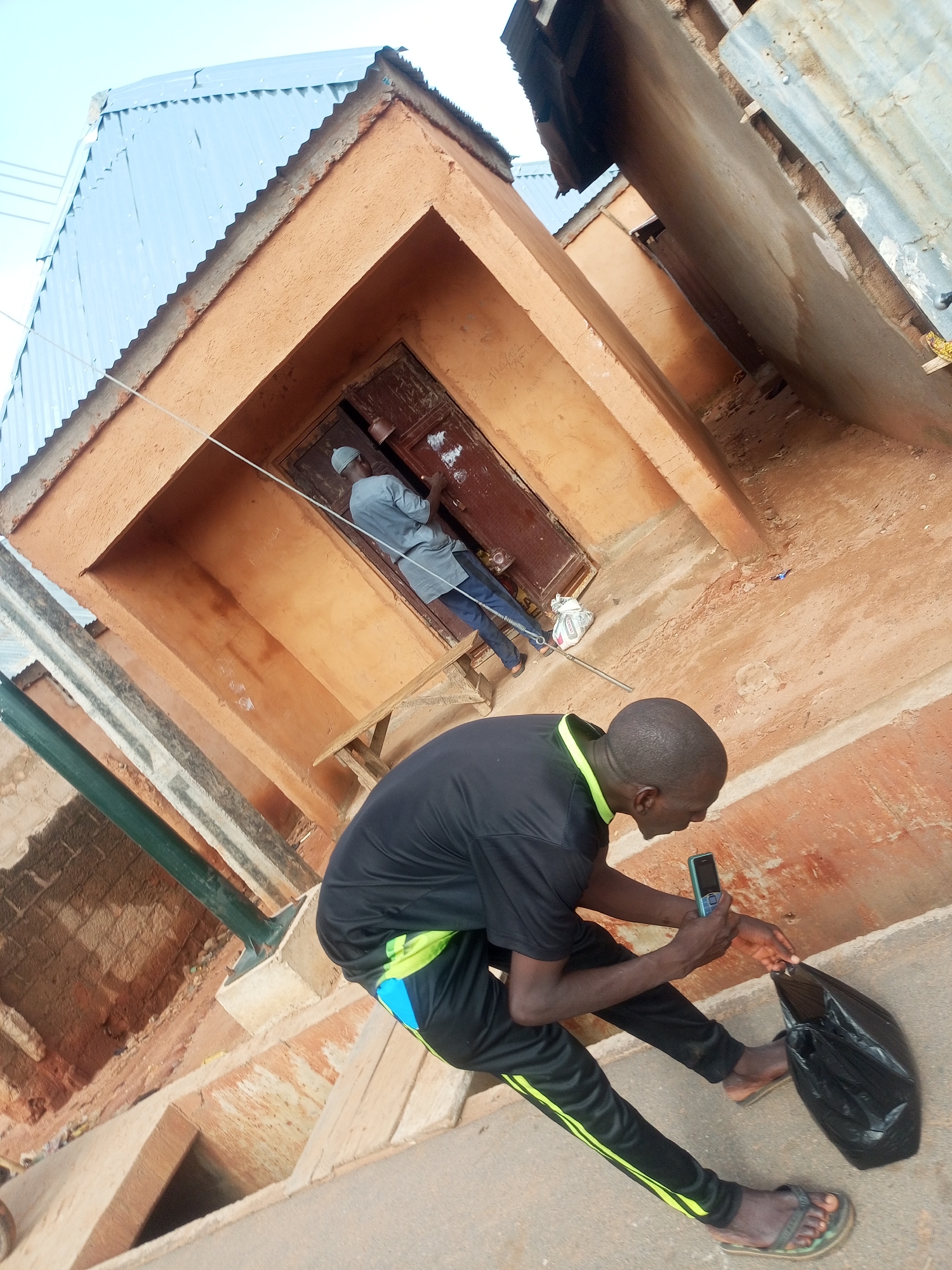 The image depicts an outdoor scene in Gombe, Nigeria. In the foreground, a man wearing a black t-shirt with a lime green collar and black track pants with lime green stripes is seated, looking at a mobile phone he is holding in his right hand. A black plastic bag rests on the ground beside him. His left hand is reaching towards the bag. He is wearing brown sandals.
In the background, a person wearing a light grey long-sleeved shirt and a white cap is standing near a wooden door. The door is set within an orange-colored building with a corrugated metal roof. The building appears to be a shop or dwelling. A wooden bench is visible beneath the doorway. The overall environment suggests a street or courtyard area.