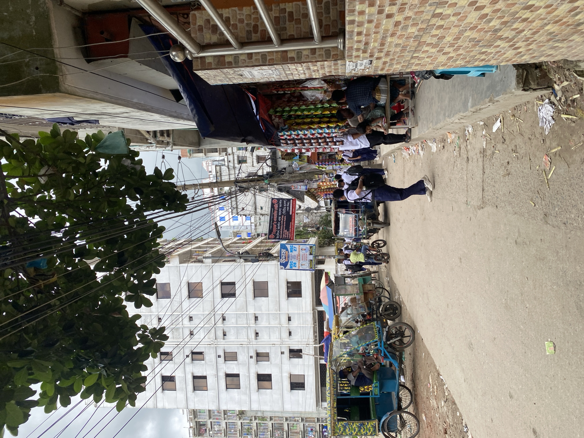 This is an eye-level, medium shot capturing a street scene in Dhaka, Bangladesh, dominated by a large white, multi-story building. The building has numerous windows with brown frames. In the foreground, a paved street is visible, with some debris scattered on the ground. To the left, a blue and green motorized rickshaw is parked, with a person sitting inside. Further down the street, several other rickshaws and carts are lined up. A group of people, many appearing to be schoolchildren in uniform, are walking or standing on the sidewalk to the right. A small shop is set up on the sidewalk, displaying various goods hanging from the ceiling. A person in a checkered shirt and a child are seated on a bench in front of the shop. Overhead, a dense network of electrical wires crisscrosses the frame, partially obscuring the sky, which appears overcast. Green foliage from a tree hangs down from the top left of the image. The overall impression is a busy urban environment with commercial activity and pedestrian traffic.