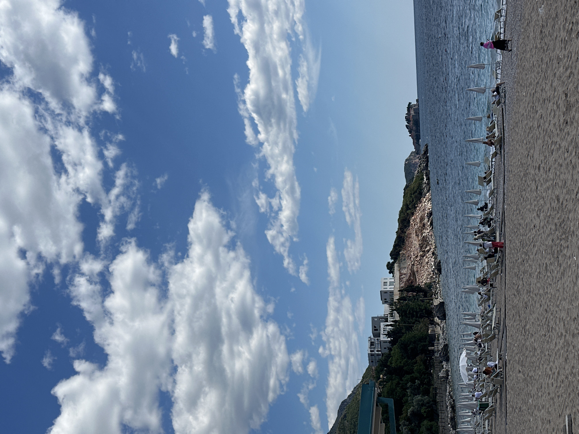 The image depicts a sun-drenched coastal scene at Kamenovo, Montenegro, on a bright day. The foreground on the right side of the frame features a sandy and pebbly beach stretching into the distance. Lined up along the shoreline are numerous white sun loungers, many accompanied by white beach umbrellas, some providing shade. Several individuals are visible relaxing on these loungers, enjoying the beach atmosphere. One person in a pink top stands near the water's edge.
To the left of the beach, the calm, blue expanse of the sea extends towards a rocky, verdant coastline. This coastline is dotted with modern, white multi-story buildings nestled among the green vegetation. Further along the coast, a distinctive rocky headland juts out into the sea, topped by what appears to be a historic or castle-like structure.
The upper and left portions of the image are dominated by a clear, bright blue sky filled with scattered, fluffy white cumulus clouds. A dark green or blue structure, possibly part of a pier or an observation deck, is partially visible in the bottom-left corner. The overall impression is one of leisure and summer vacation at a scenic Mediterranean location. No visible text is present.