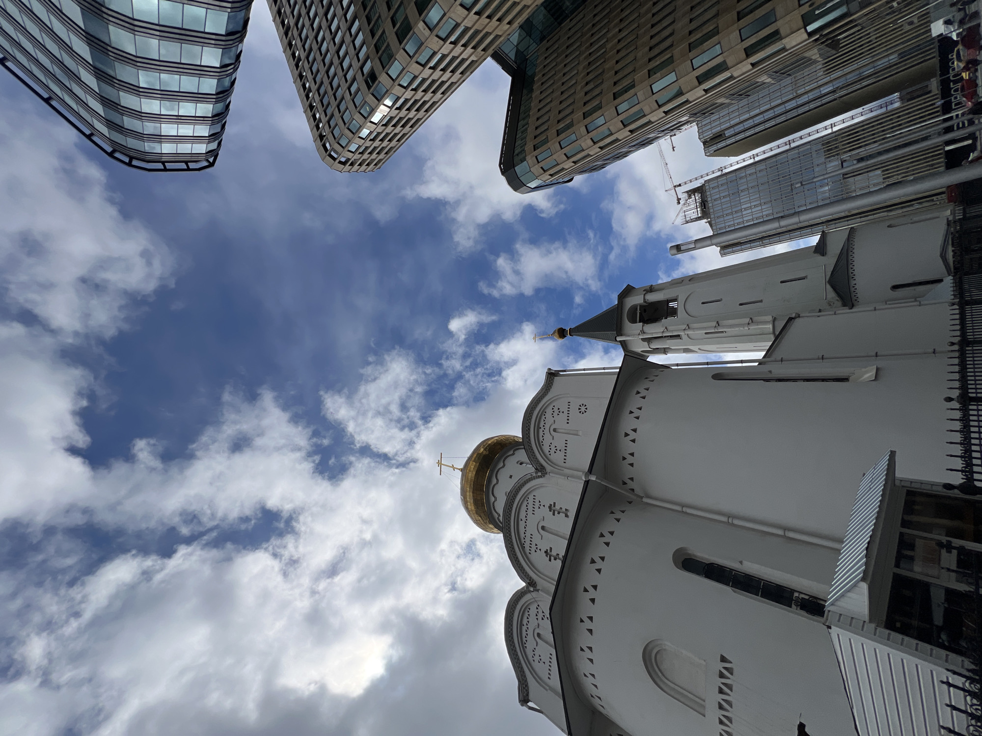 The image, taken from a low angle looking up, shows a convergence of architectural styles against a backdrop of a cloudy blue sky in Moscow, Russia. To the right is a white Orthodox church, easily identifiable by its golden domes and intricate decorations on its walls, next to modern tall buildings. In the top left and middle sections, contemporary glass and concrete high-rise buildings loom over the church, creating a stark contrast between old and new architecture. A construction crane is visible in the background of the modern buildings. The cloudy sky provides a dynamic and visually interesting background. No people are visible in the frame.