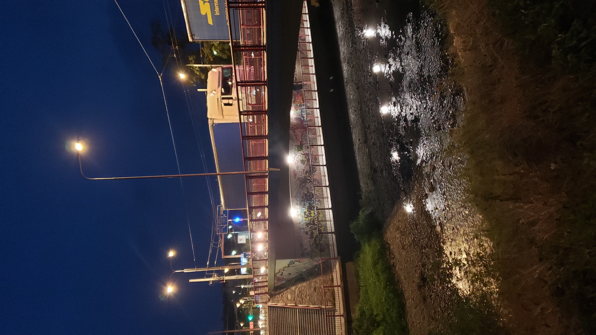 This image depicts a nighttime scene in the city of Prešov, Slovakia. A bridge with red railings spans over a shallow, rocky riverbed or drainage channel. The bridge is illuminated by streetlights and lights from vehicles. A white truck is visible on the bridge, and a blue truck with a DHL logo is partially visible on the right side. The underside of the bridge is covered in graffiti. On the left side of the image, there are stone steps leading up to the bridge. The riverbed reflects the lights from above, creating a shimmering effect. The surrounding environment appears to be urban, with trees and utility poles visible. The sky is dark blue, indicating it is nighttime.