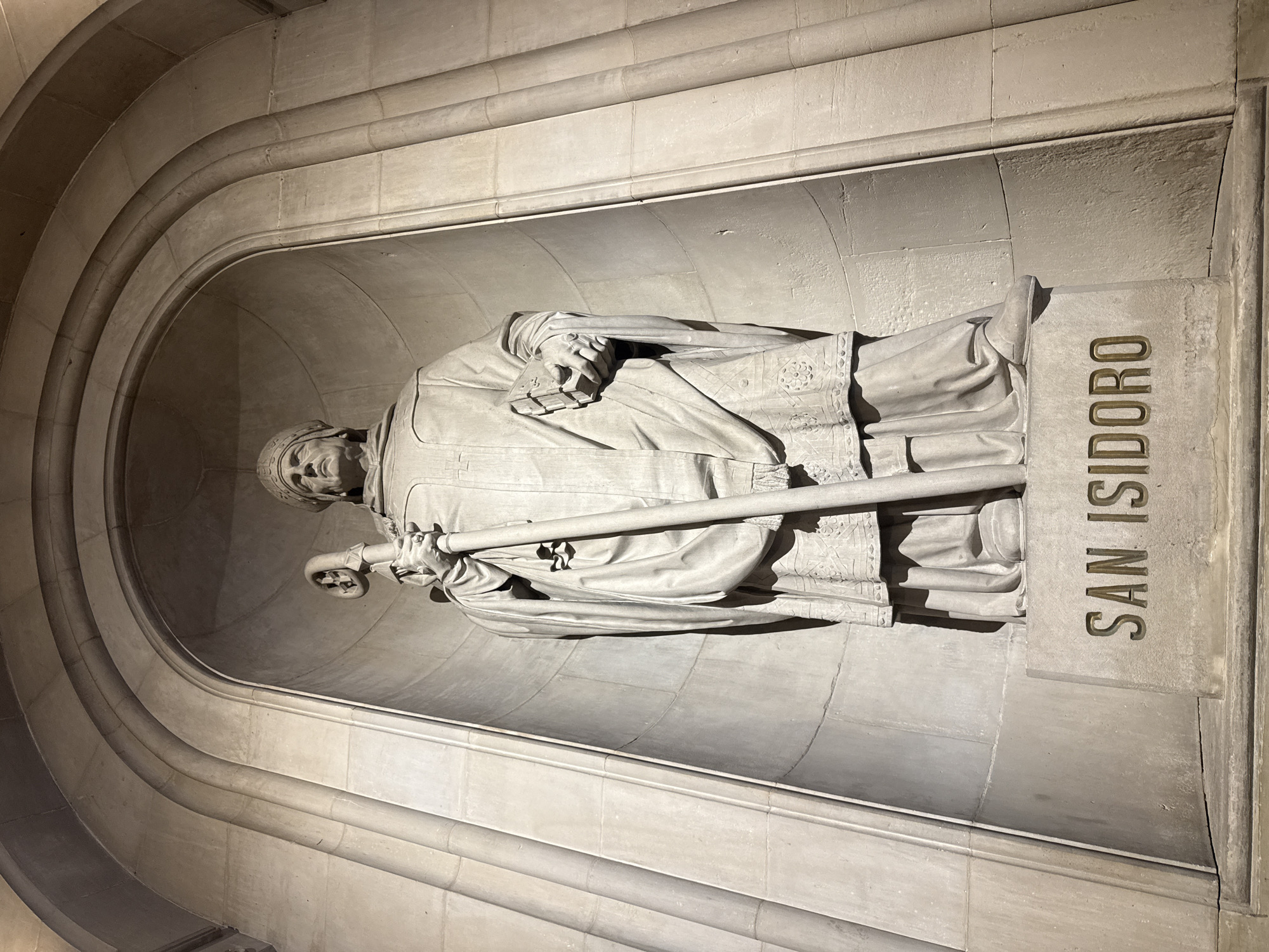 The image depicts a stone sculpture of San Isidoro (Saint Isidore) located in a niche within a stone wall or structure. The sculpture, carved from light-colored stone, portrays San Isidoro standing and holding a staff. He is wearing a clerical robe or gown. The name "SAN ISIDORO" is inscribed in gold letters on a stone plaque to the right of the sculpture. The sculpture is housed in a curved niche. The location is the Basilica of the Sagrada Familia in Barcelona, Spain.
